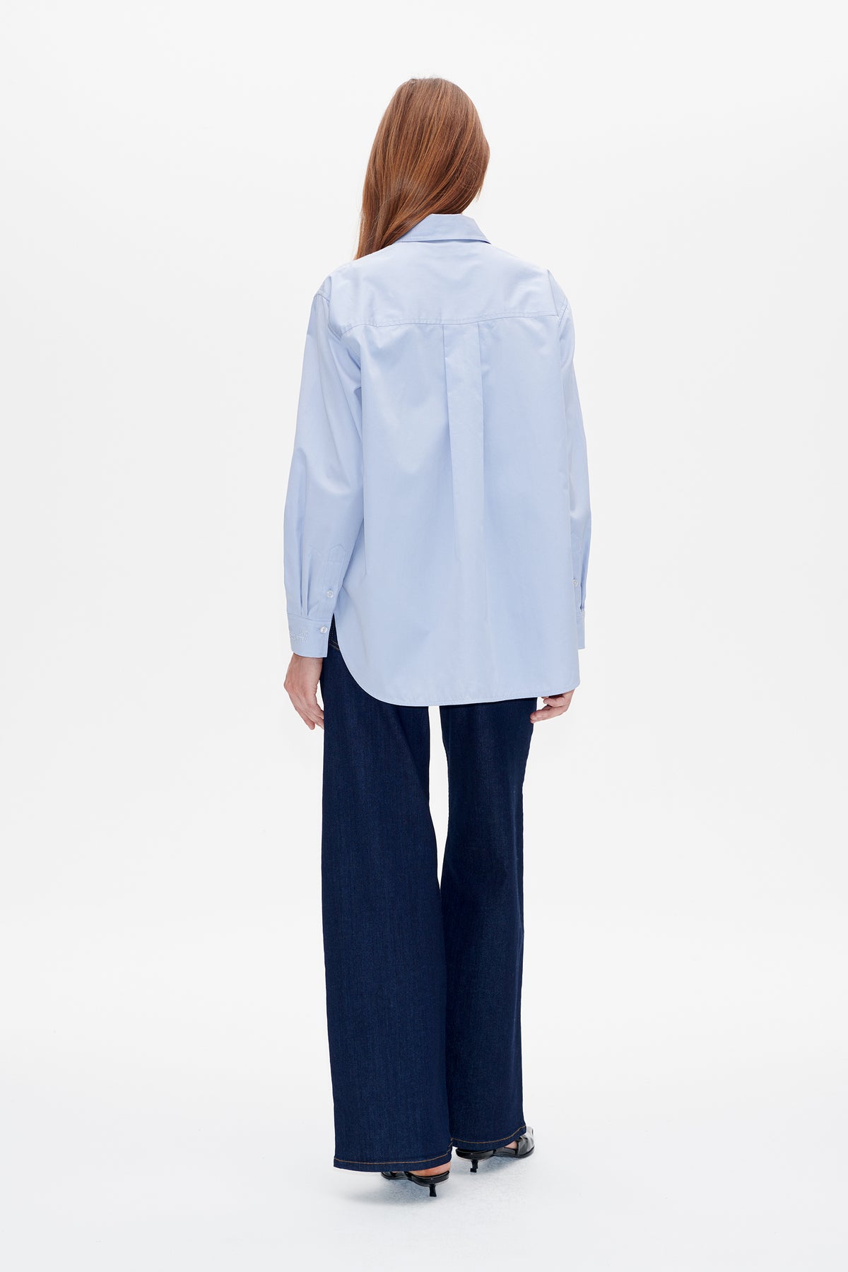 Back view of a model wearing a pale blue long-sleeved shirt with a yoke seam and centre pleat, paired with dark wide-leg jeans.