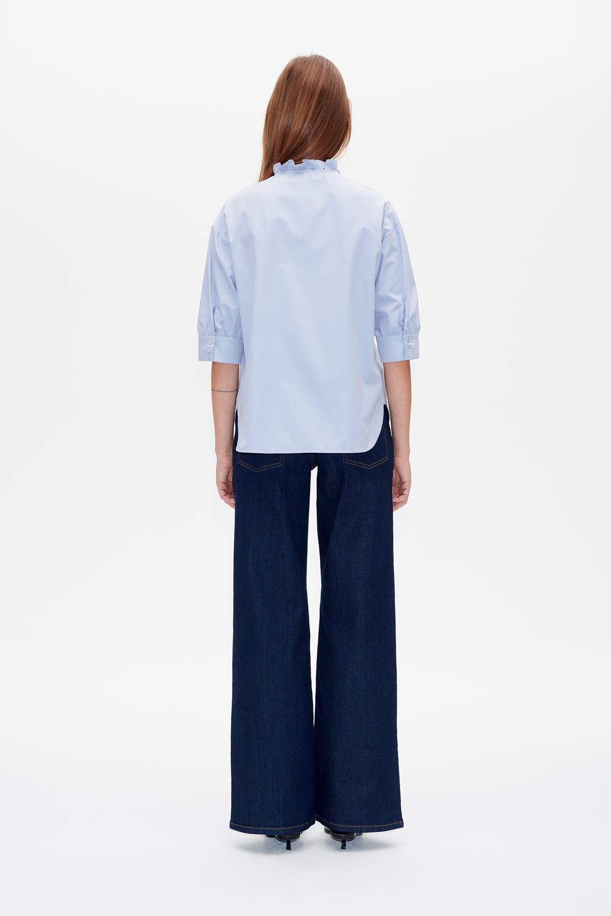 Back view of a pale blue blouse with a ruffled collar, three-quarter sleeves and buttoned cuffs, worn with dark wide-leg jeans.