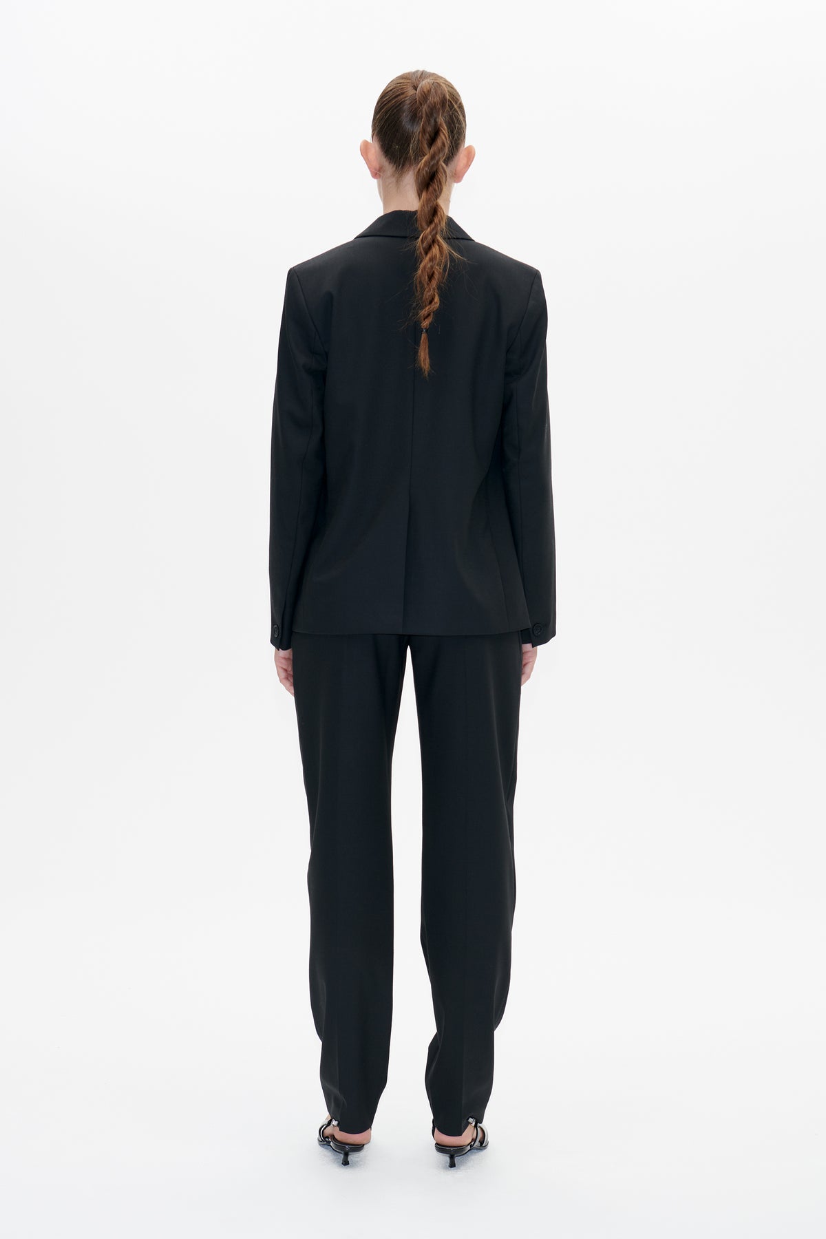 Back view of a model wearing a black tailored blazer with matching straight-leg trousers and low-heeled sandals.