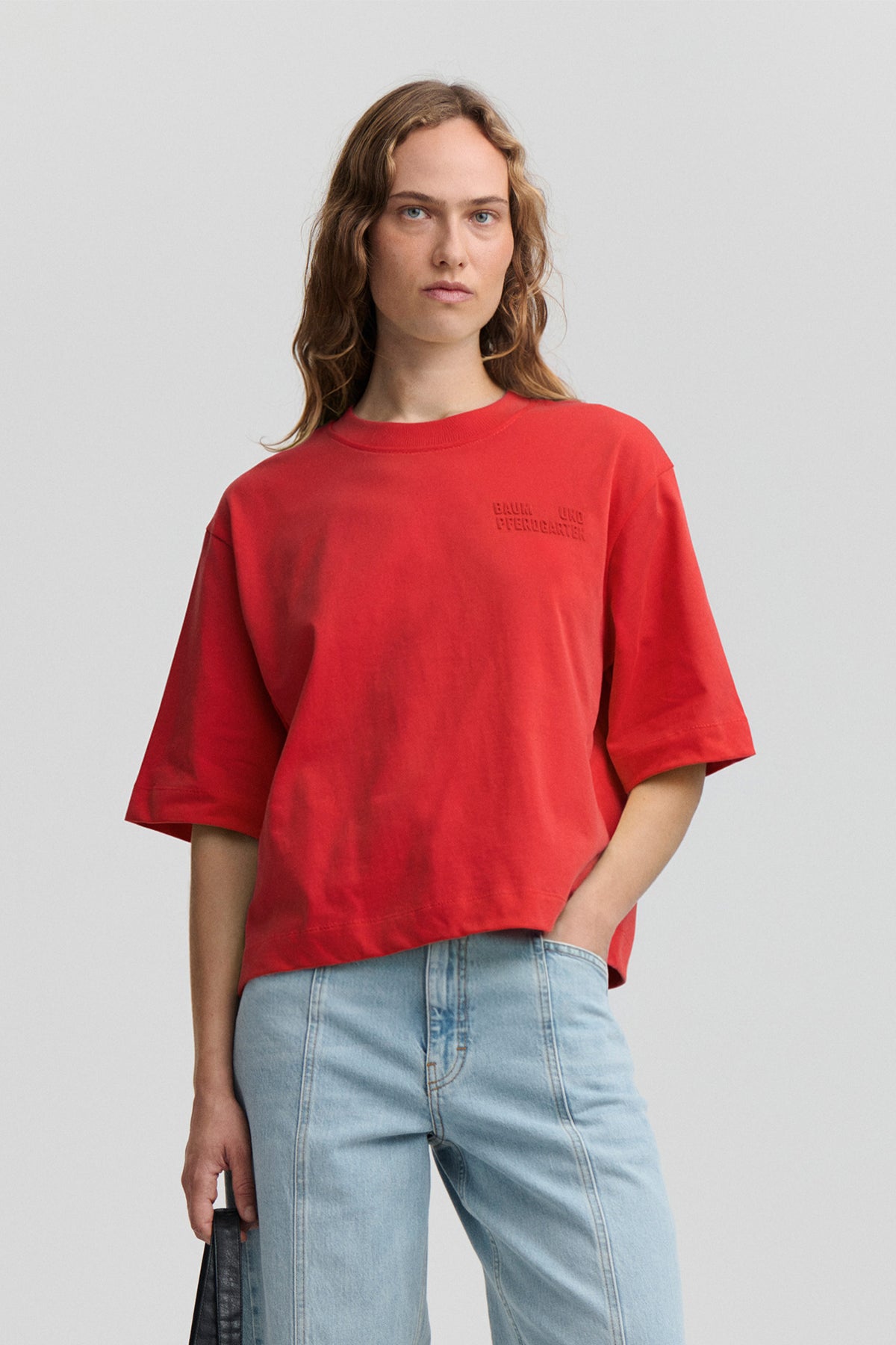 A woman modeling an oversized red short-sleeve t-shirt with a tonal embroidered logo on the left chest, paired with light blue jeans, against a solid light gray background.