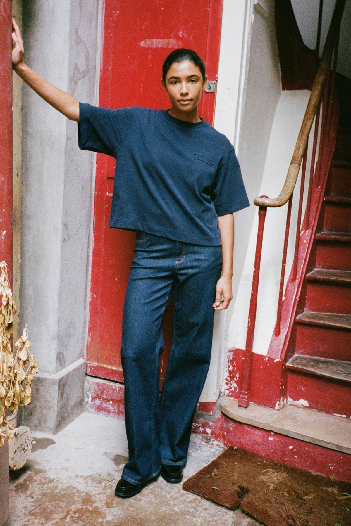 A model poses in a navy oversized short-sleeve T-shirt with subtle chest lettering and dark wide-leg jeans, framed by a red doorway.