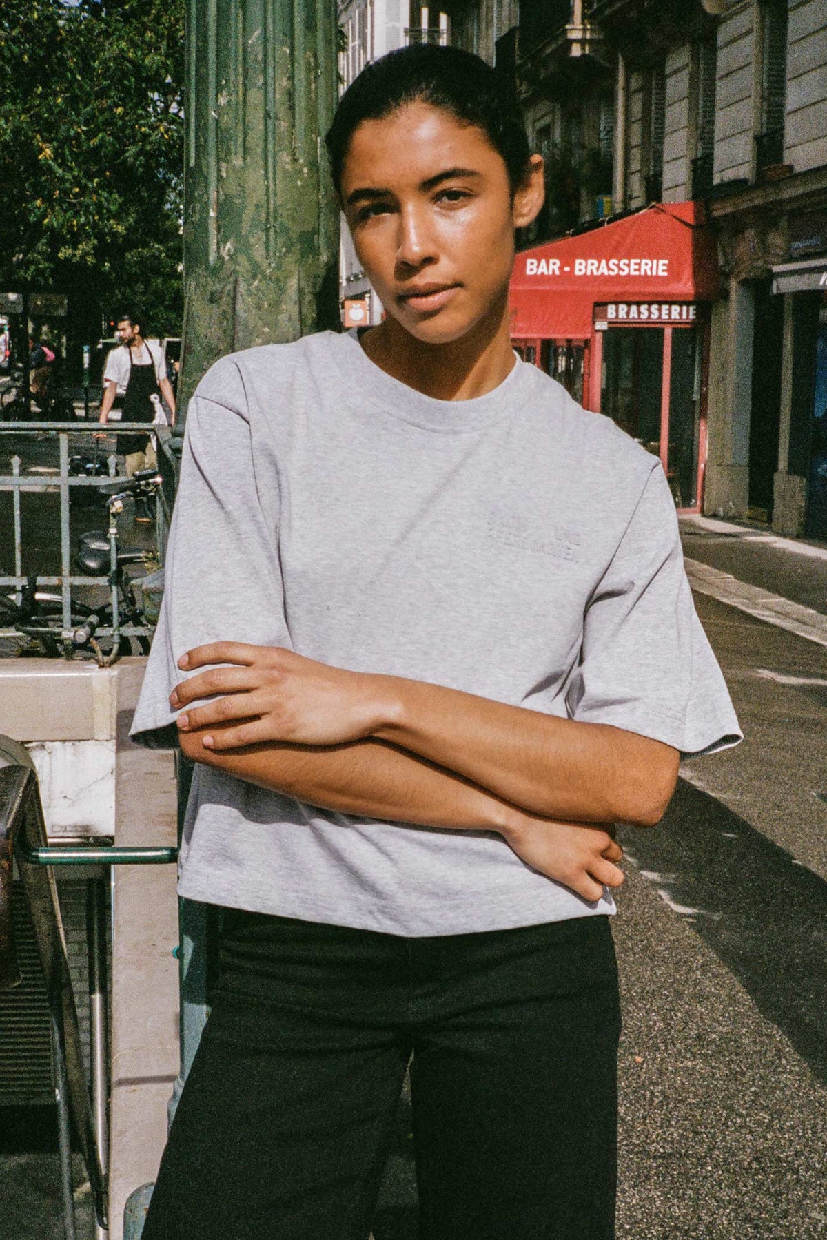 Street-style campaign portrait of a person in a plain light grey short-sleeved T-shirt and black trousers, standing with arms crossed.