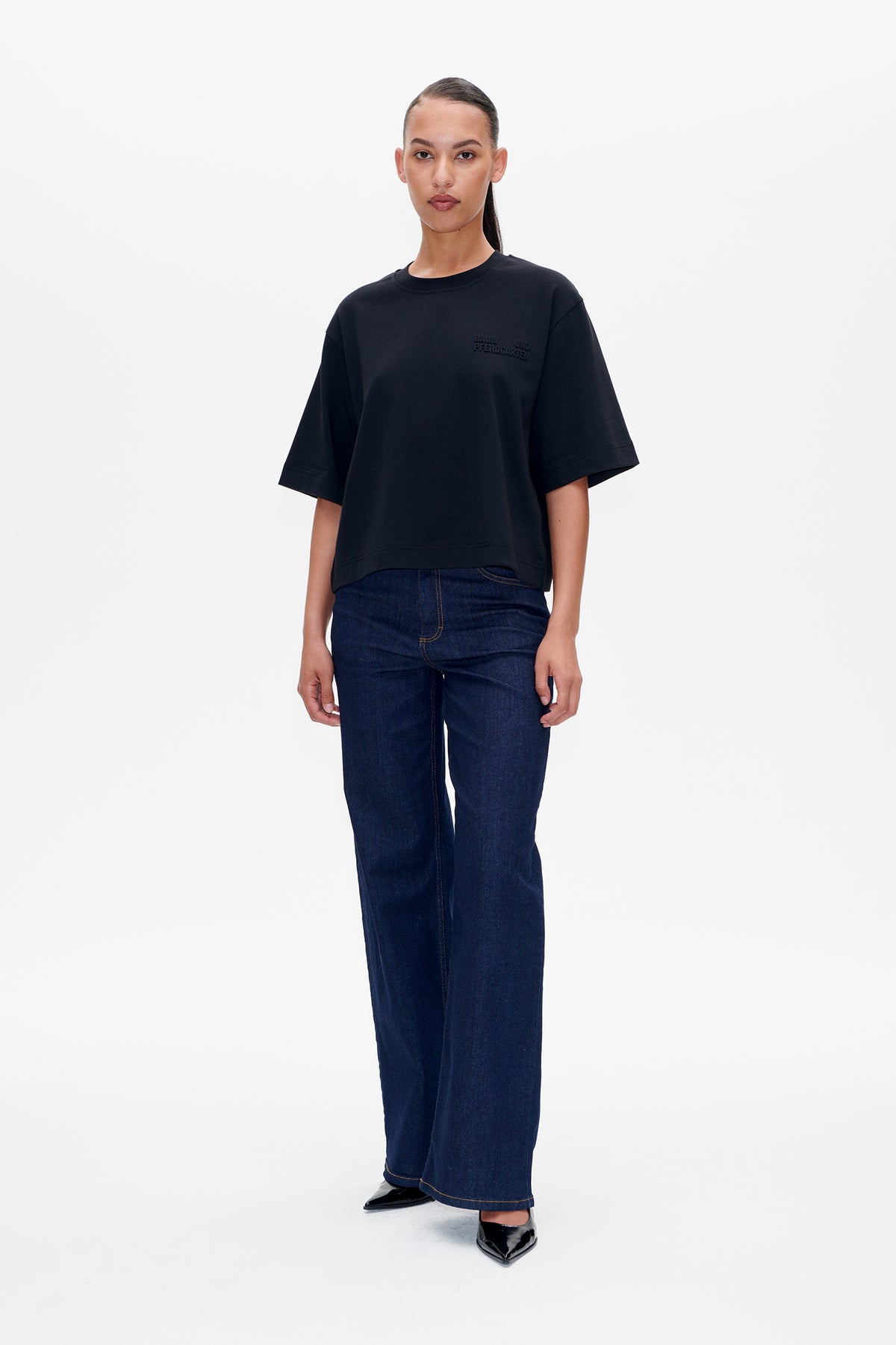 Model draagt een zwarte oversized T-shirt met korte mouwen en een klein borstlogo, gecombineerd met donkerblauwe wijde jeans. Vooraanzicht.