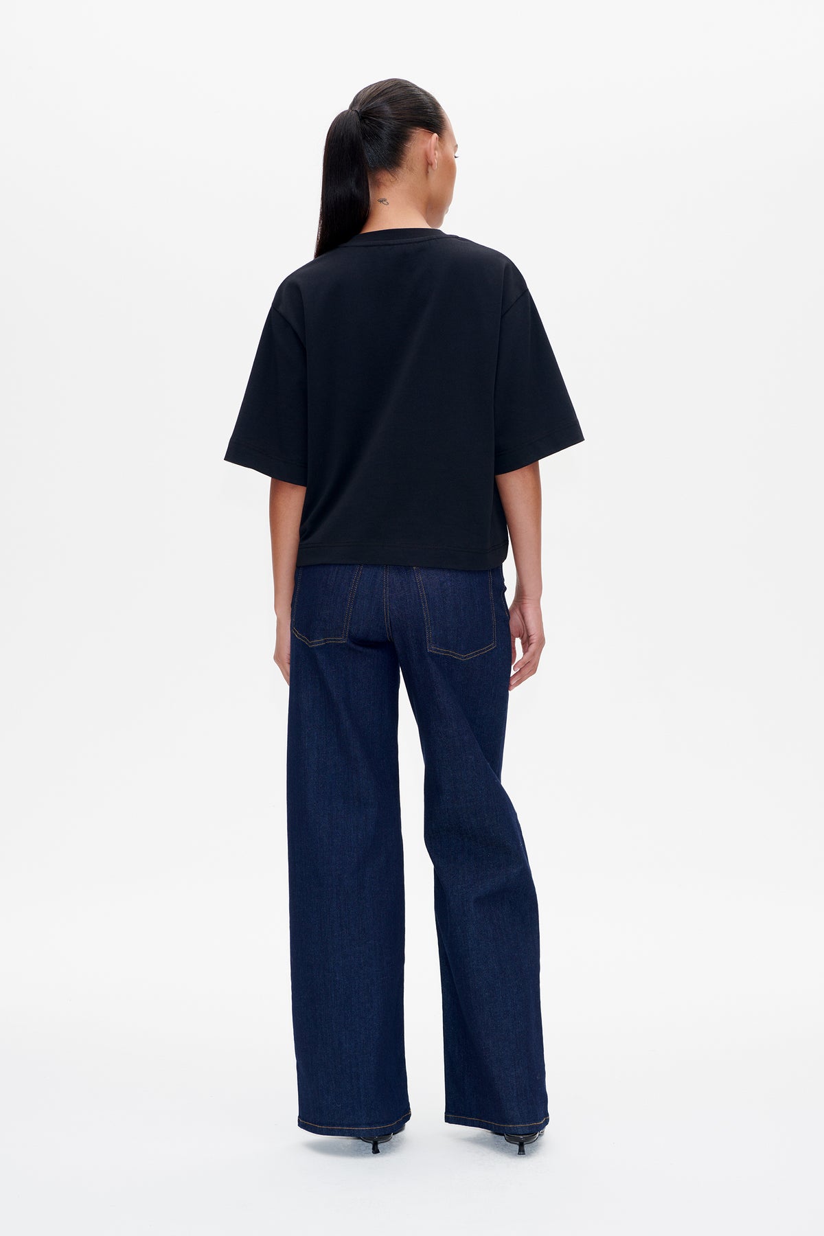 Achteraanzicht van een model dat een effen zwart T-shirt met korte mouwen draagt en donkerblauwe wijde jeans.
