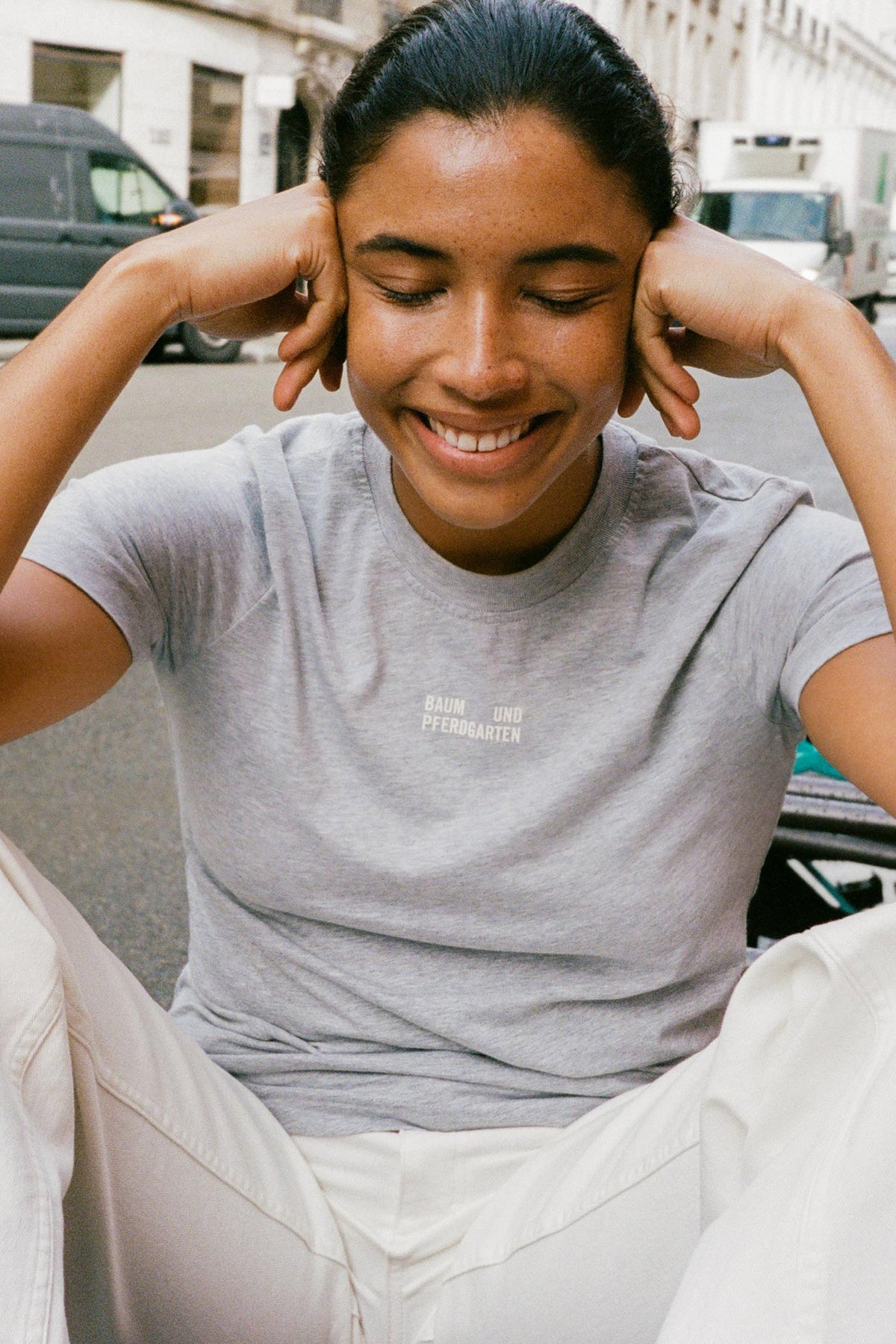 Campaign close-up of a smiling model in a light grey T-shirt with a small white Baum und Pferdgarten chest logo.