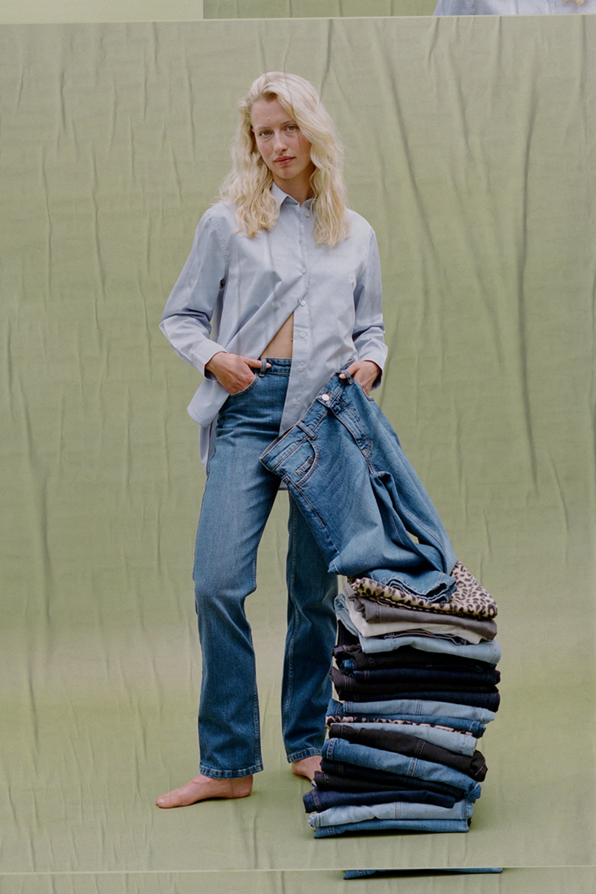 Model in a pale blue button-up shirt and blue jeans holds denim beside folded clothes against a pale green backdrop.
