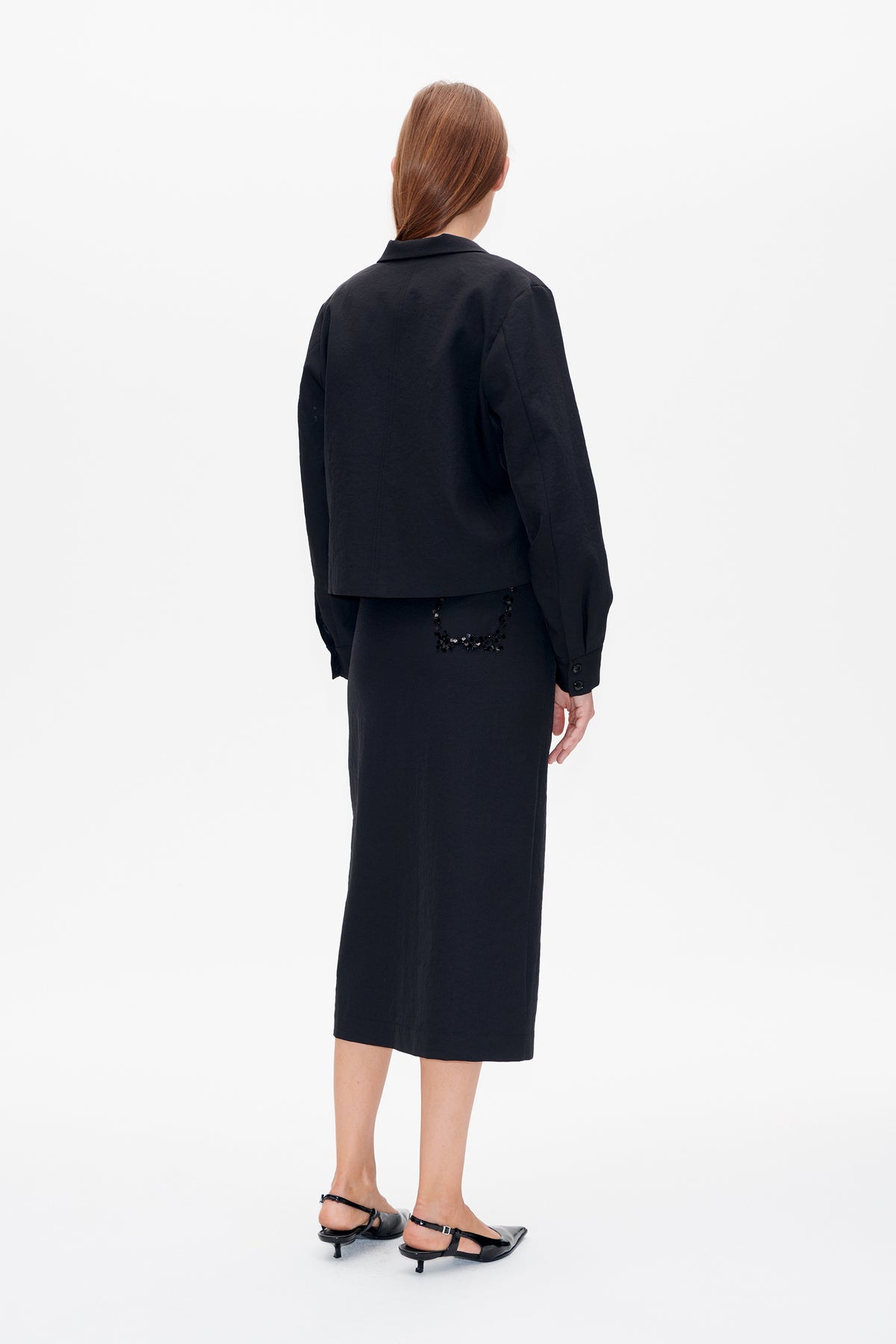 Back view of a model in a black collared jacket and long black skirt with a square sequin embellishment at the back.