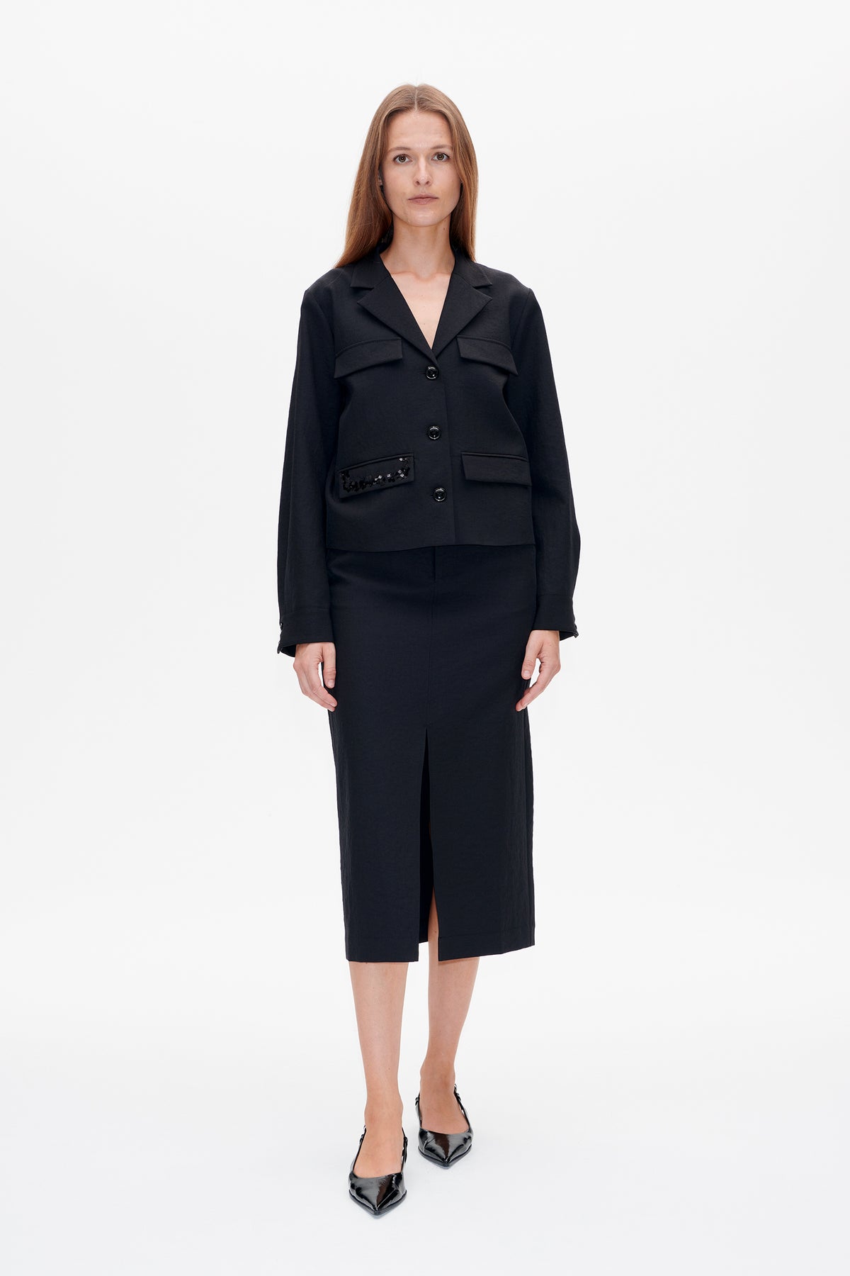 Front view of a model wearing a black button-front jacket with flap pockets and a sequined detail, paired with a long black slit skirt.