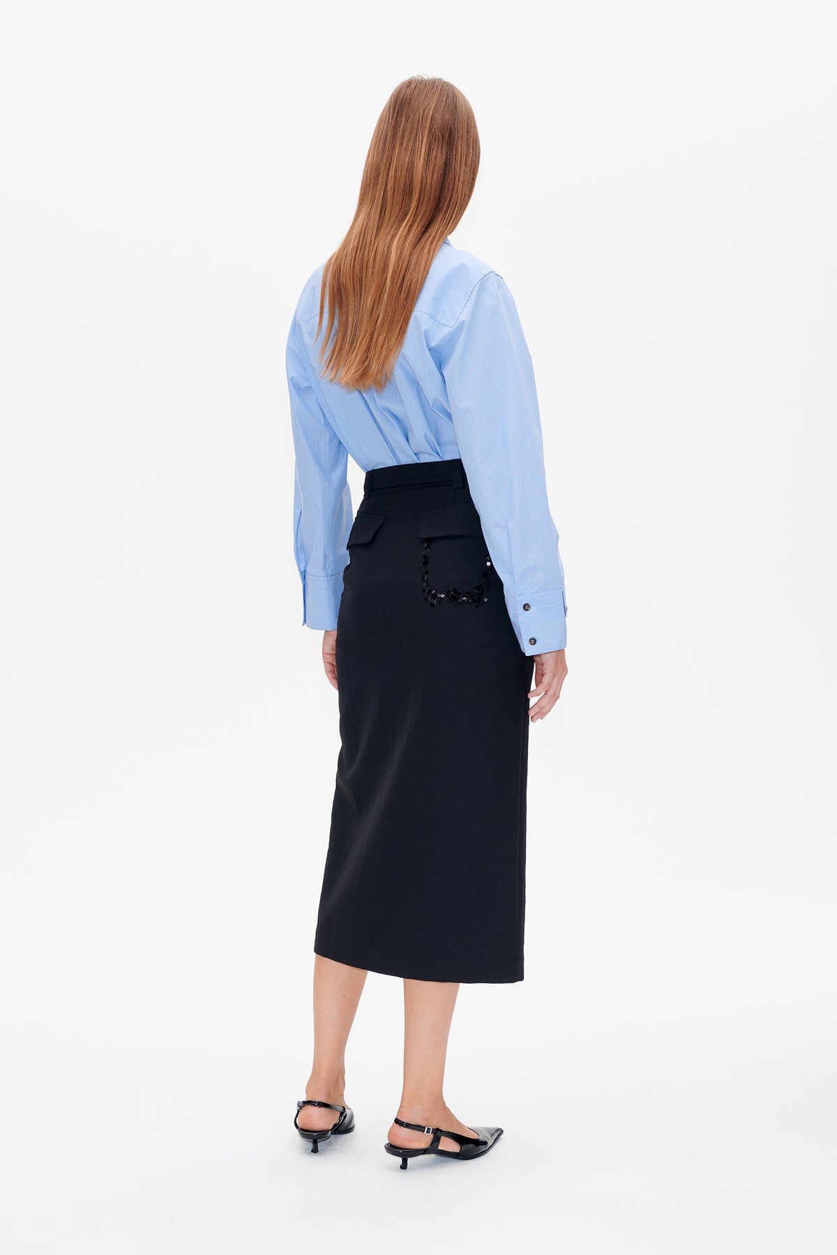 Back view of a model wearing a pale blue pinstriped shirt and black midi skirt with embellished rear pocket flaps.