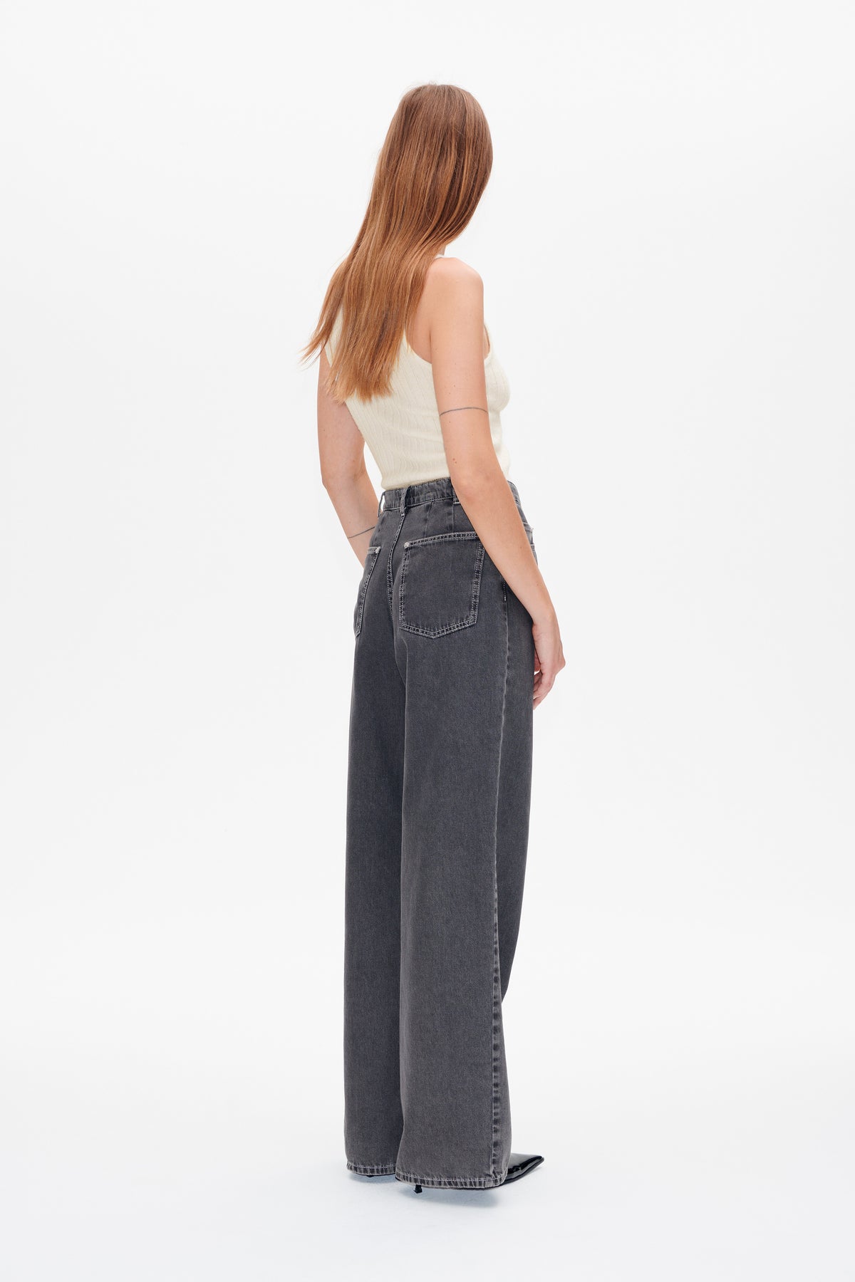 Achteraanzicht van een model dat een crèmekleurige, mouwloze ribgebreide top draagt en wijde antracietgrijze jeans met contrasterende stiksels.