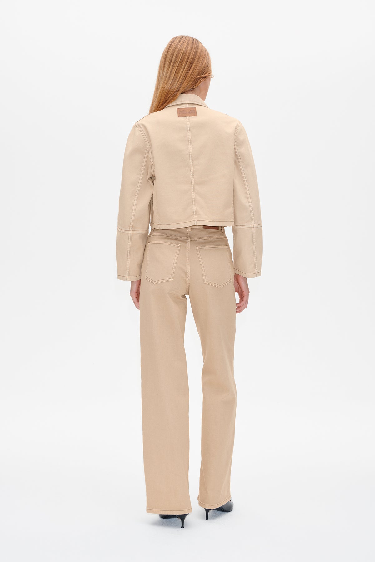 Rückenansicht eines Models, das eine kurze, beige Jacke mit Nahtdetails und eine passende, weit geschnittene Hose trägt.