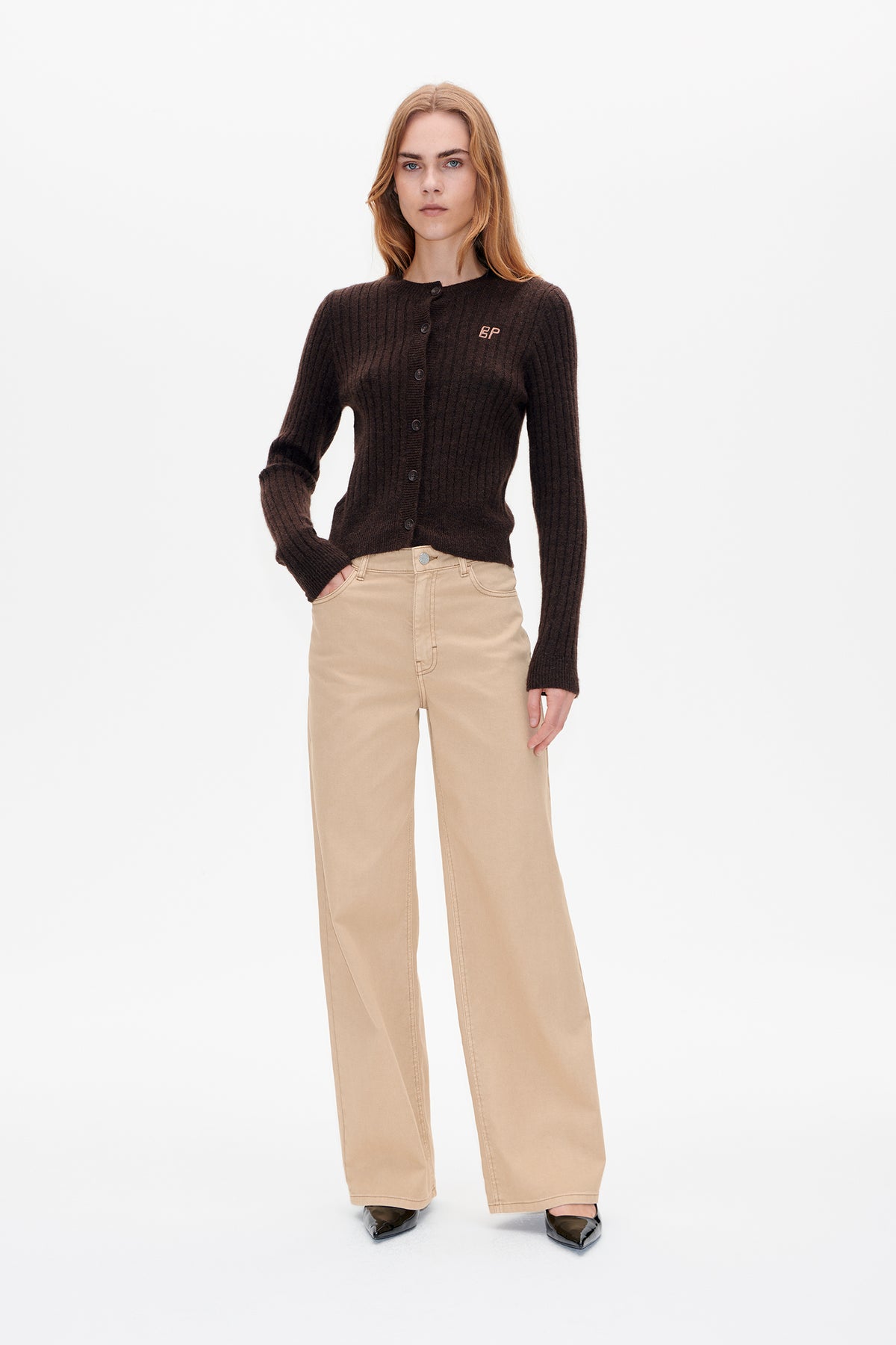 Front view of a model wearing a dark brown ribbed button-front cardigan with a small chest logo, beige wide-leg trousers and black pointed shoes.