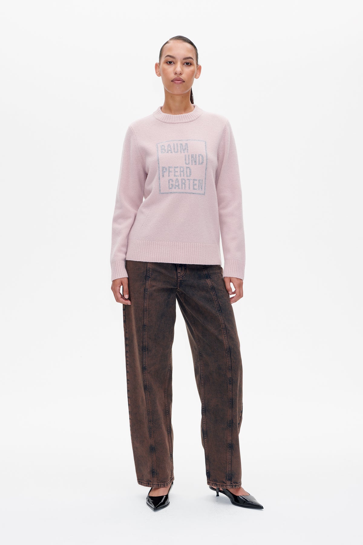 Front view of a model wearing a pale pink crew-neck sweater with a large Baum und Pferdgarten chest logo, brown jeans and black pointed heels.