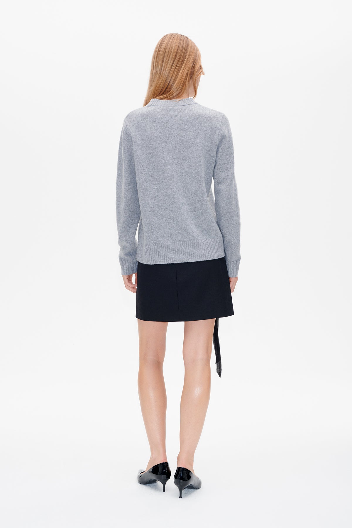 Back view of a model wearing a light grey knit sweater with ribbed cuffs and hem, paired with a black mini skirt.
