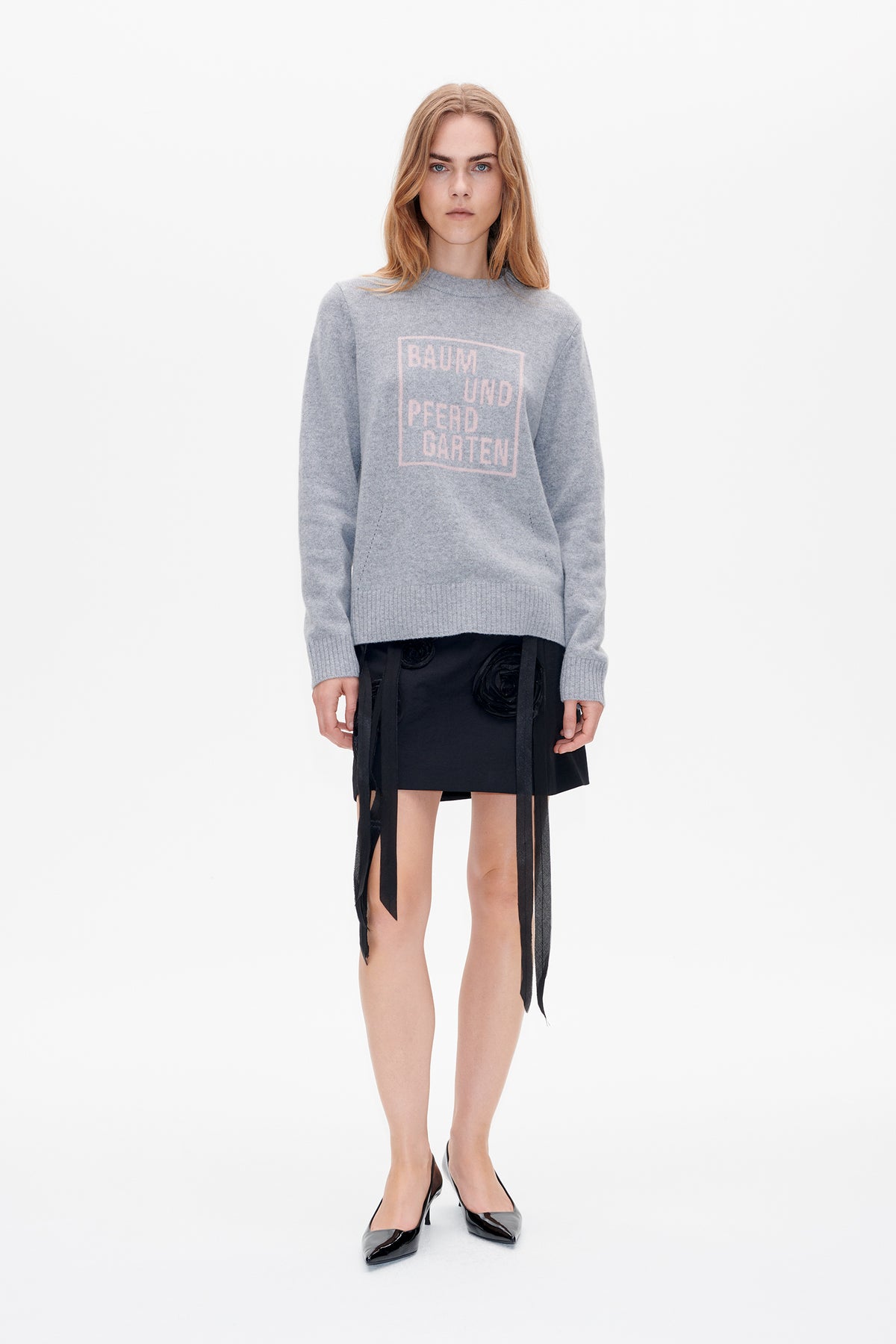 Front view of a model wearing a light grey crew-neck sweater with pale pink Baum und Pferdgarten lettering, a black skirt with sheer hanging panels, and pointed heels.