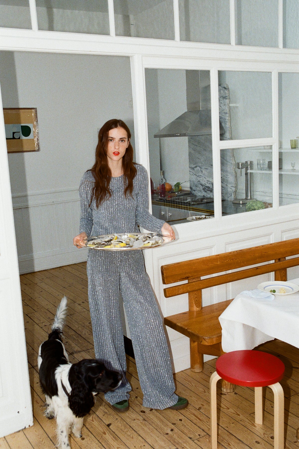 A model holds a platter in a kitchen, wearing a blue-grey long-sleeved top and wide-leg trousers with a small white print; a dog stands nearby.