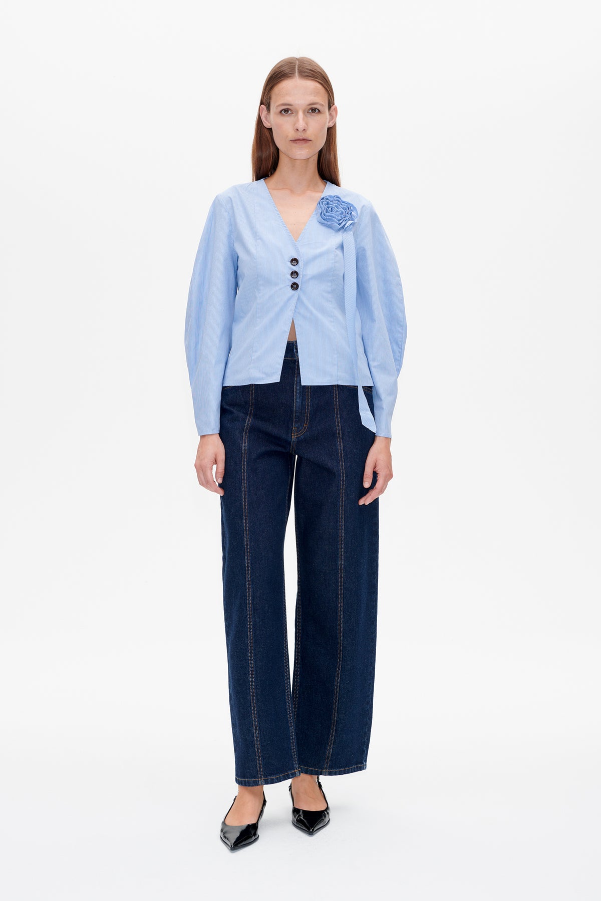 Front view of a light blue pinstriped blouse with a deep V-neck, three buttons and a fabric flower detail, paired with dark wide-leg jeans.