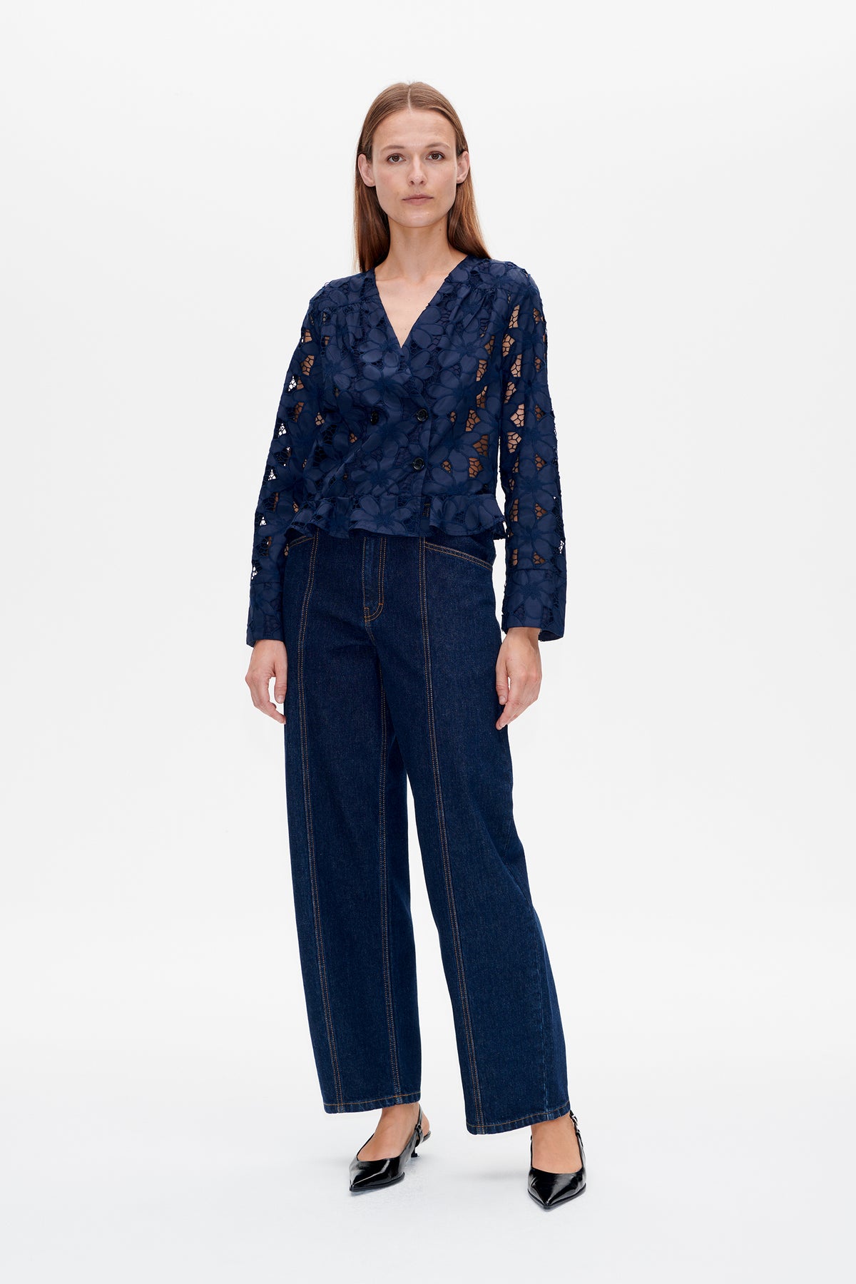 Vorderansicht eines Models in einer marineblauen Jacke mit floralen Cutwork-Details, V-Ausschnitt und doppelter Knopfleiste, kombiniert mit dunklen Jeans mit weitem Bein.