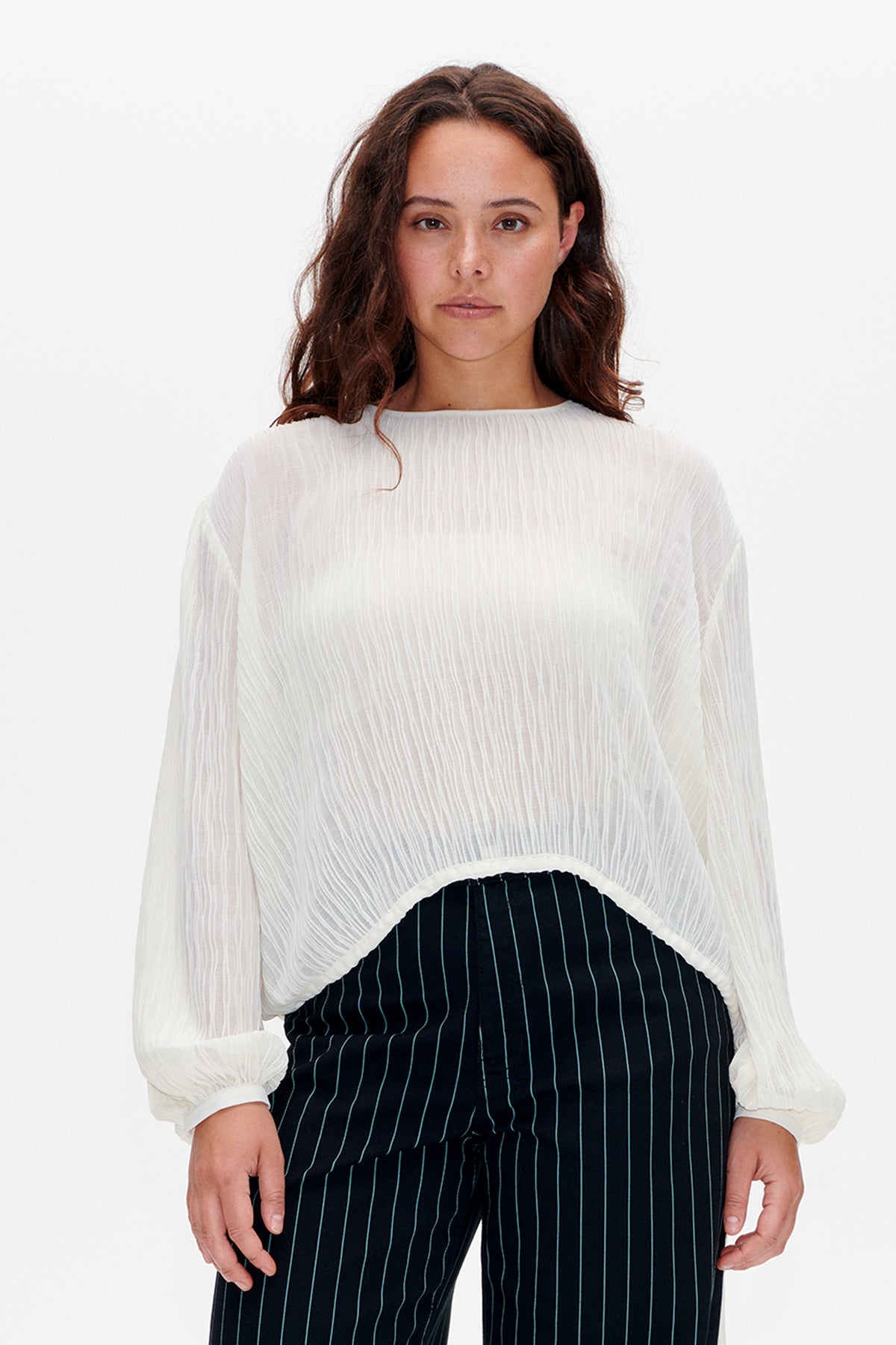 Front view of a sheer white blouse with a gathered neckline, textured vertical stripes and voluminous sleeves, worn over dark pinstriped trousers.
