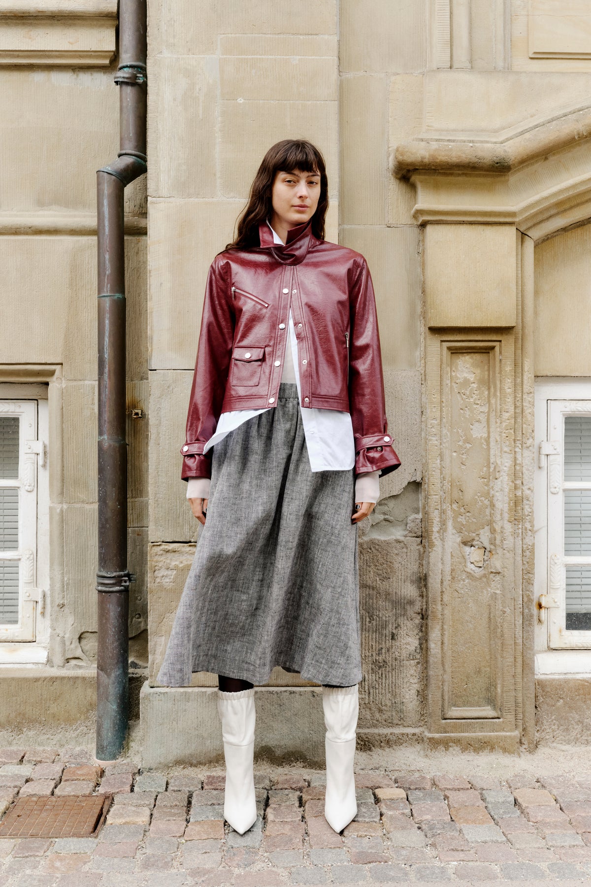 Model wearing a burgundy glossy jacket with zip pockets over a white shirt and grey herringbone skirt, standing against a stone facade.
