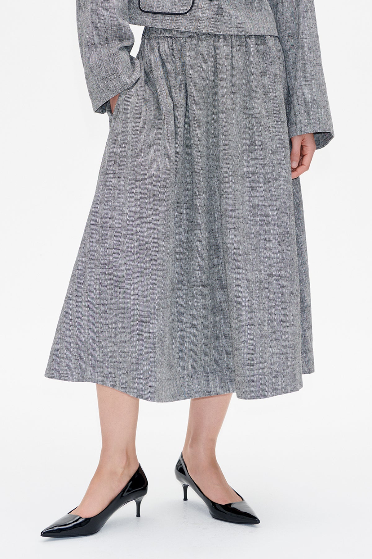 Close front view of a grey herringbone-patterned dress with a gathered waist and black-piped flap pocket, styled with black pointed heels.
