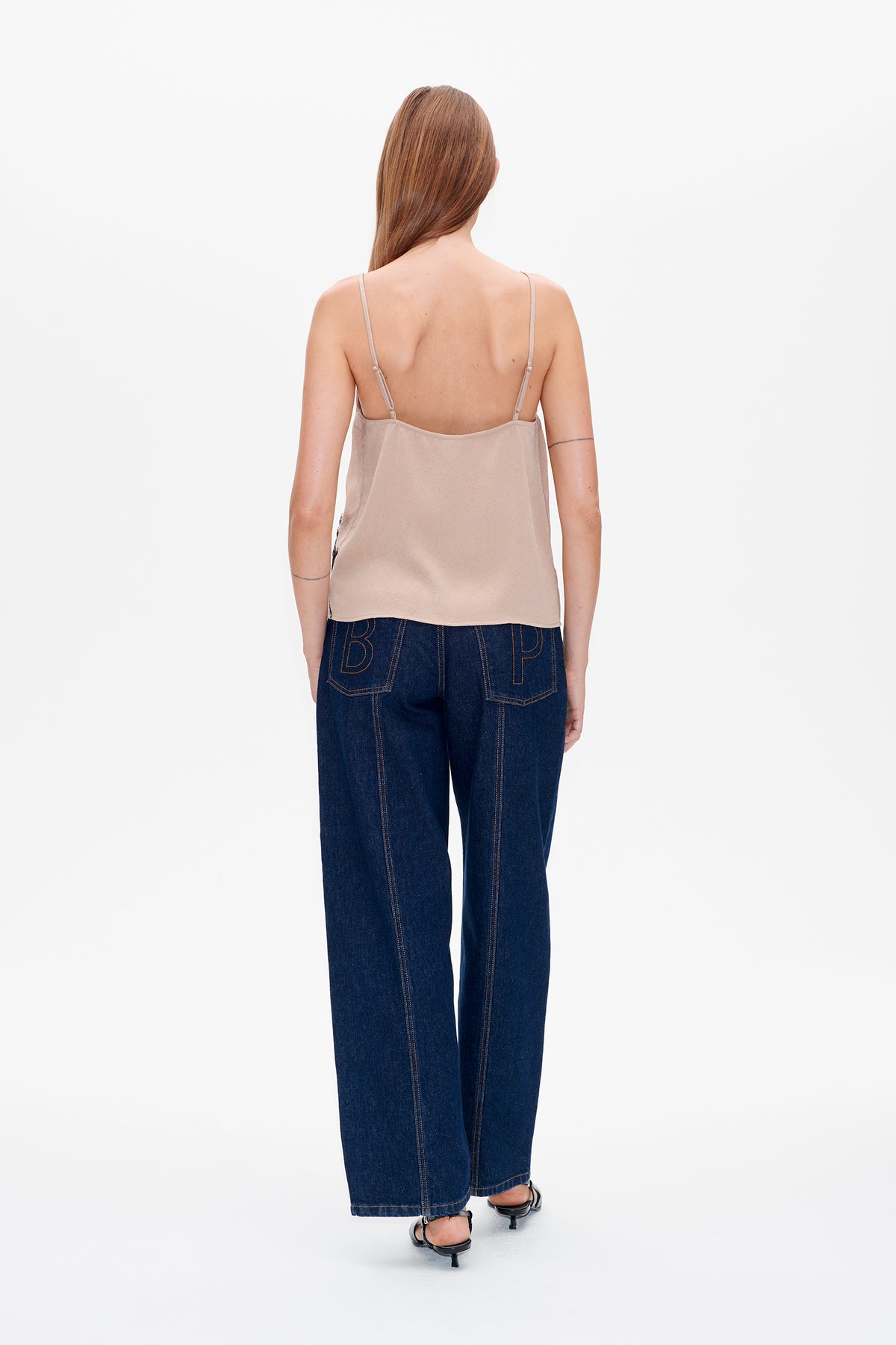 Achteraanzicht van een model in een beige cami met smalle bandjes en een wijdvallende jeans in donker indigo met grote omlijnde letters op de achterzakken.