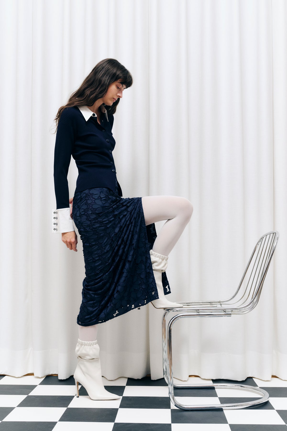 Campaign portrait of a seated model in a navy ribbed button cardigan with a white collar and a textured navy skirt.