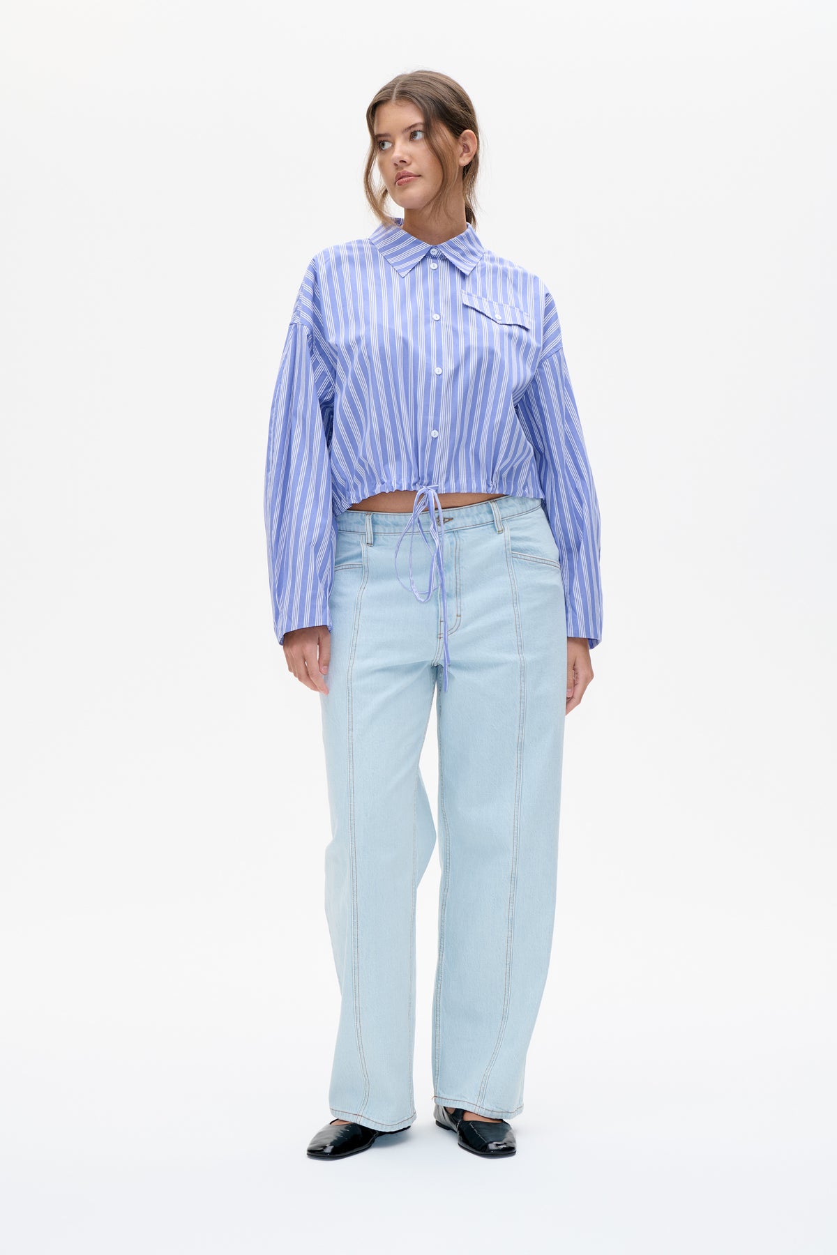 Front view of a model wearing a blue-and-white striped cropped button-up shirt with a chest pocket and drawstring hem, paired with light-wash jeans.