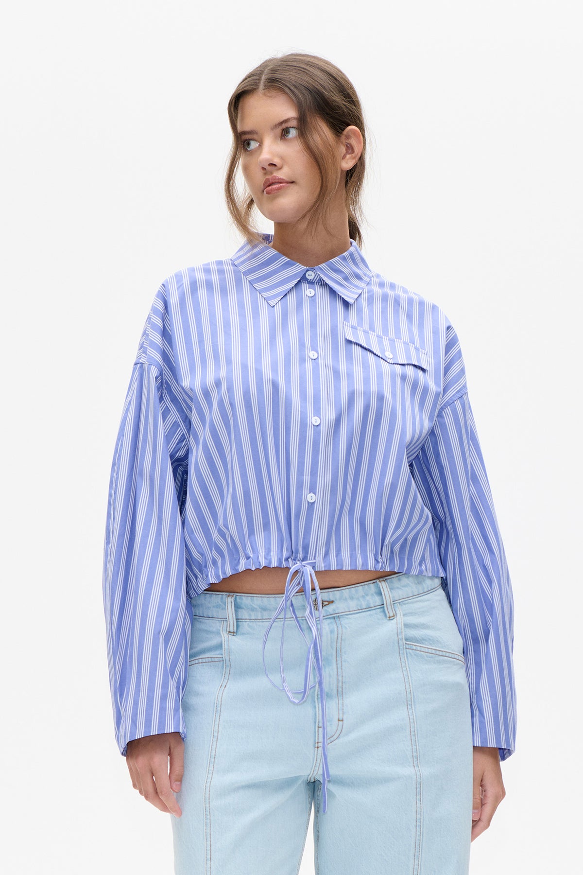 Front view of a cropped blue shirt with white stripes, pointed collar, button front, chest pocket and a tie-drawstring hem.