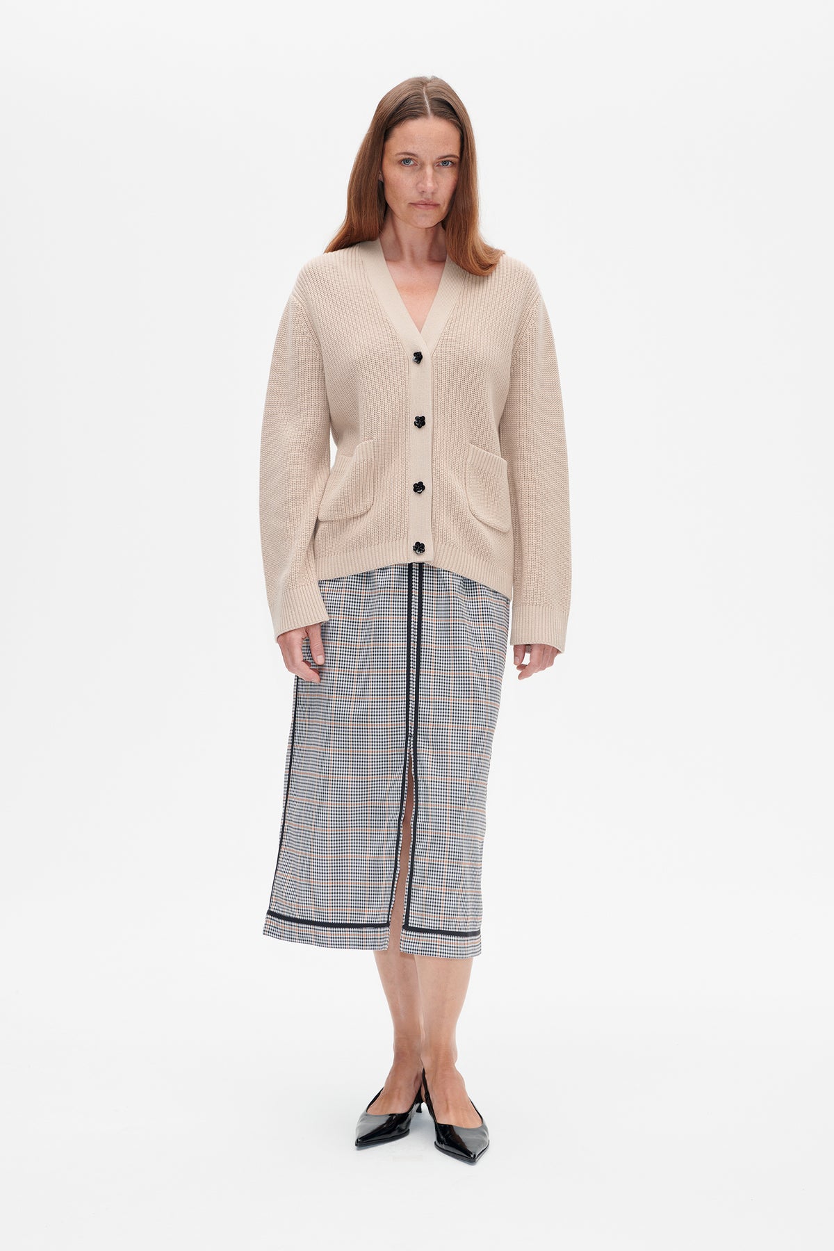 Front view of a model wearing a cream rib-knit cardigan with black flower-shaped buttons and patch pockets, over a checked skirt.