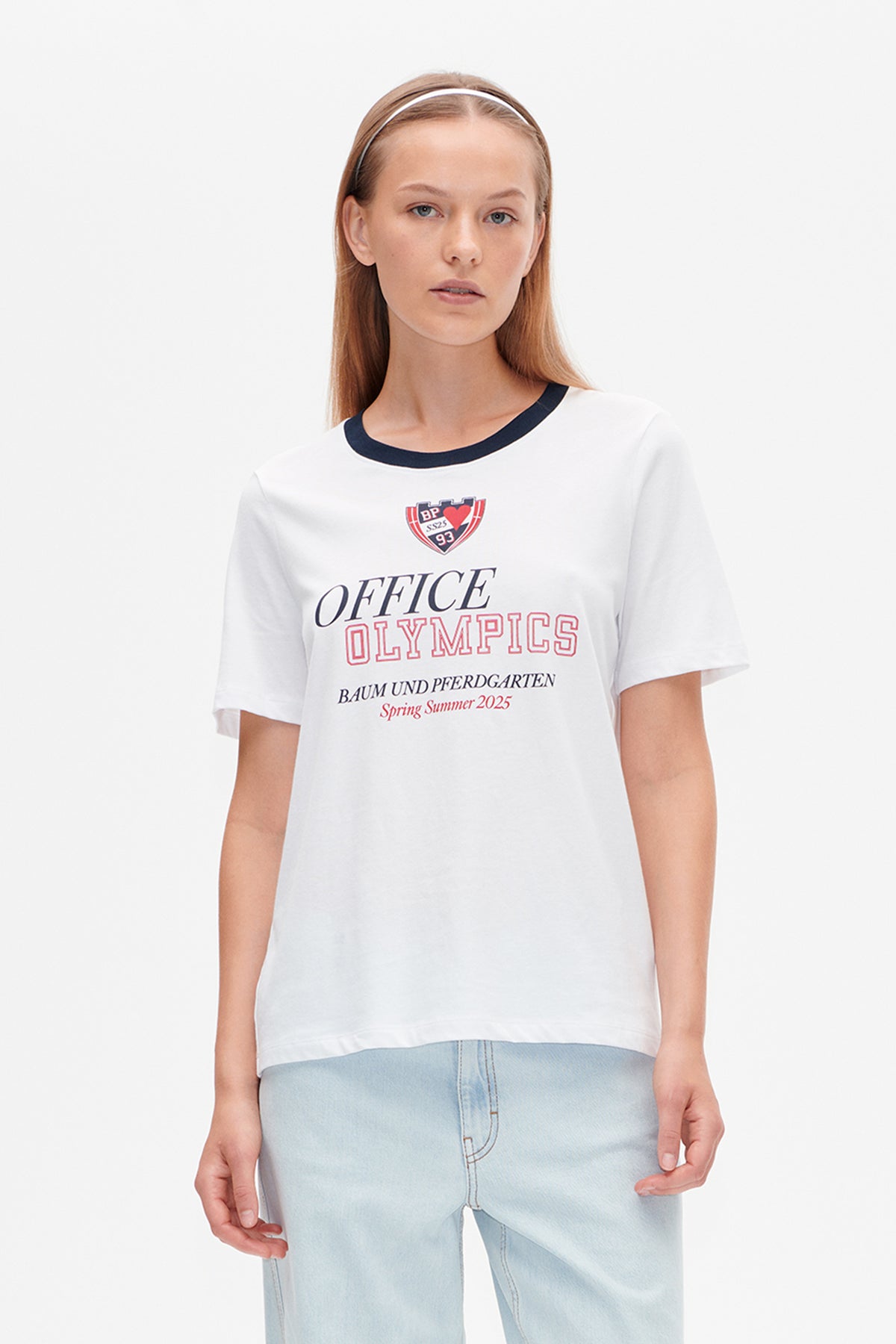 Vue de face d'un t-shirt blanc à manches courtes, col bleu marine et motif sur la poitrine « Office Olympics » rouge et bleu marine.