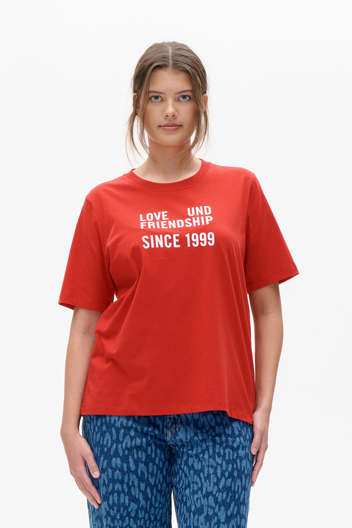 Vooraanzicht van een model dat een rood T-shirt met korte mouwen draagt, bedrukt met “Love und Friendship Since 1999”, en blauwe jeans met patroon.