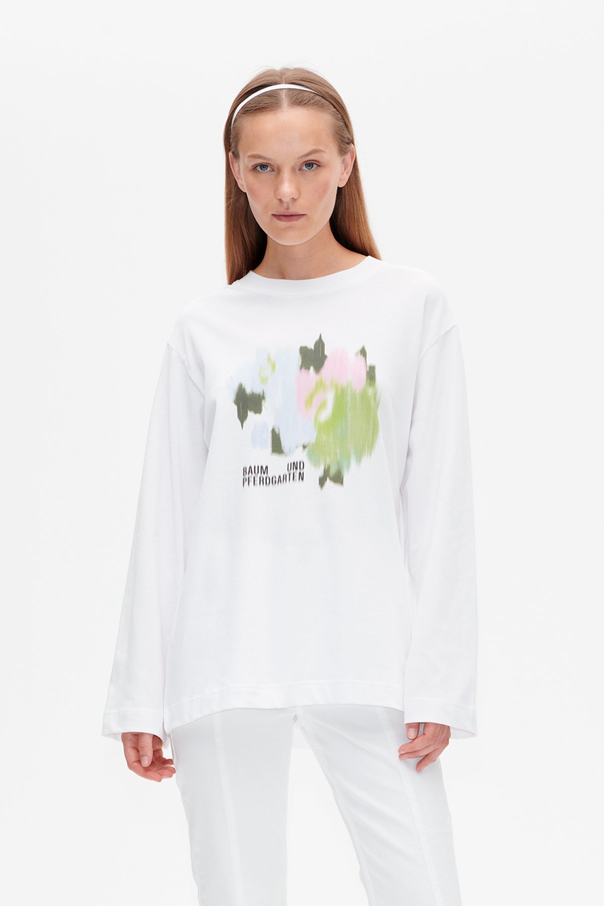 Model wearing a white long-sleeve sweatshirt with an abstract pastel graphic and Baum und Pferdgarten lettering on the front.