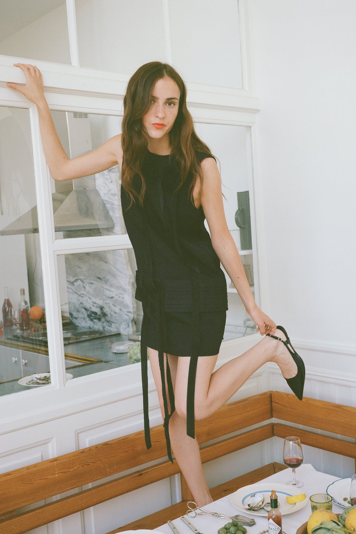 Model poses beside a dining table in a black sleeveless mini dress with long hanging ties and black heels.