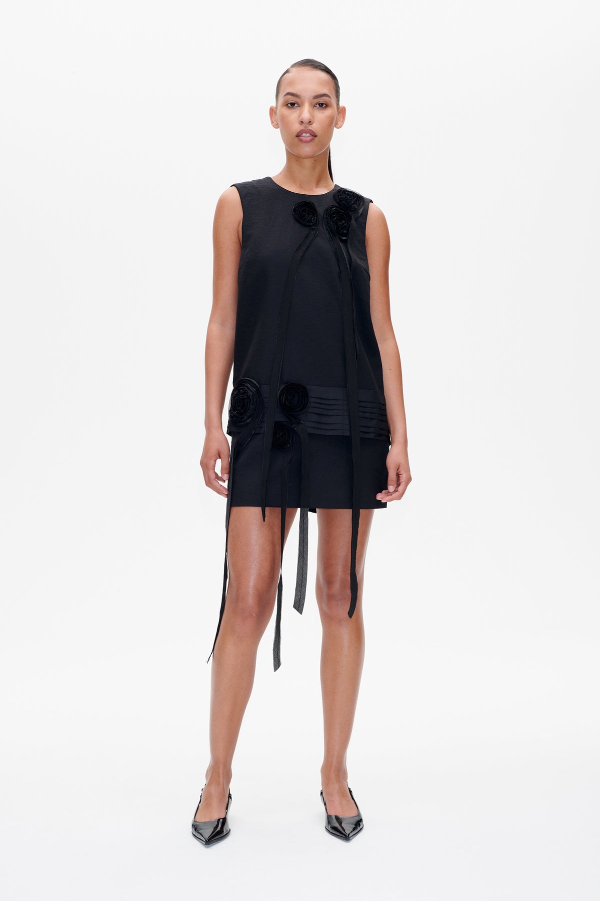 Front view of a black sleeveless mini dress with fabric rosettes, long hanging ties and pleated detail at the hem.