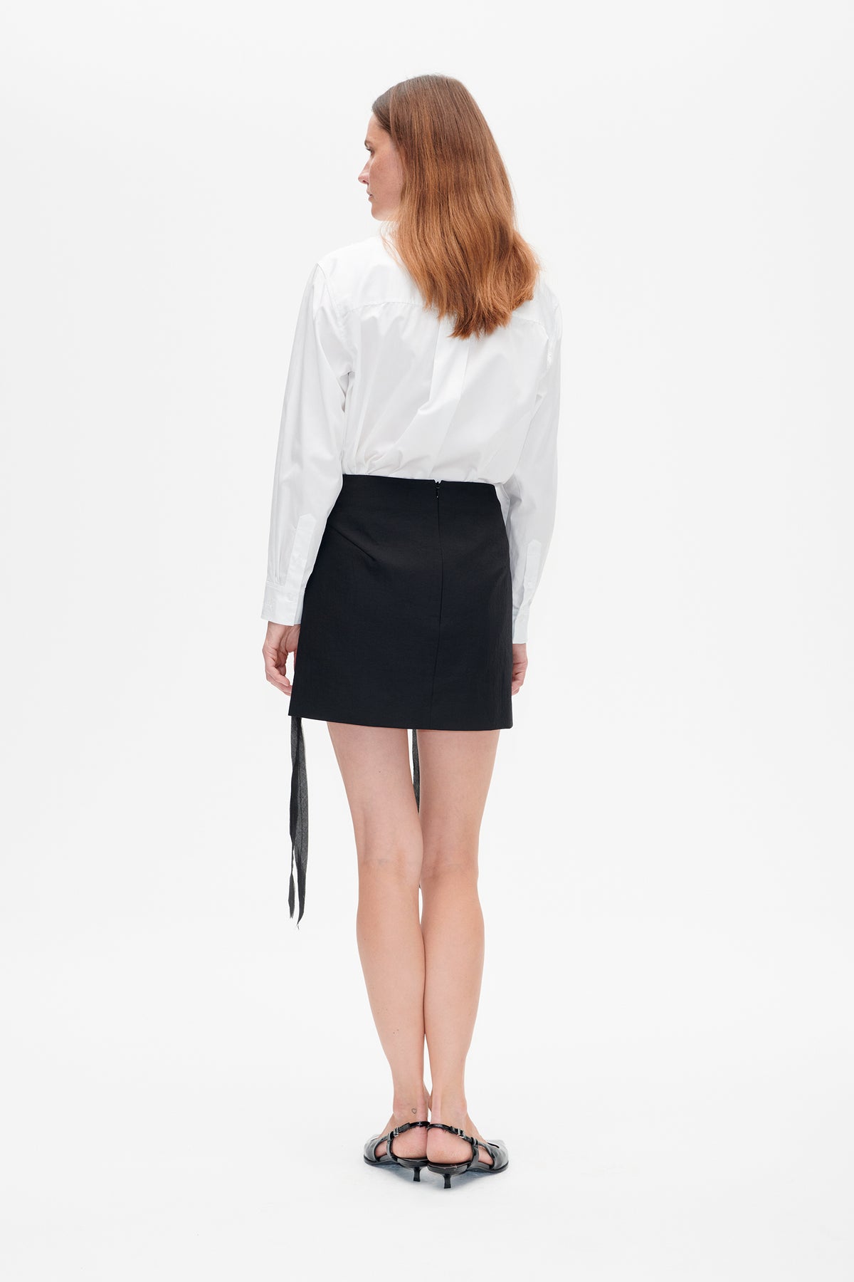Back view of a model wearing a white long-sleeved shirt tucked into a black mini skirt with long hanging ties.