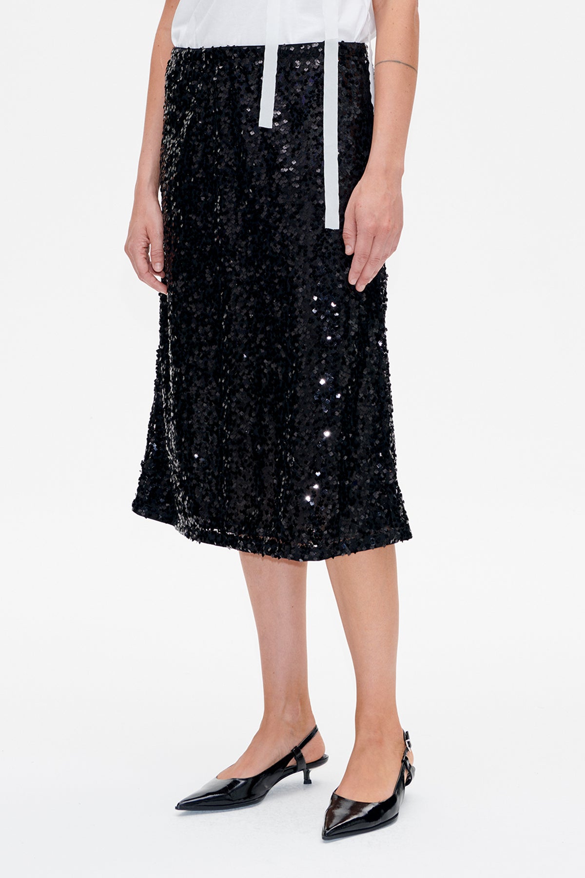 Front detail of a black sequined skirt with a white sleeveless top and long white ties, styled with pointed black slingback heels.