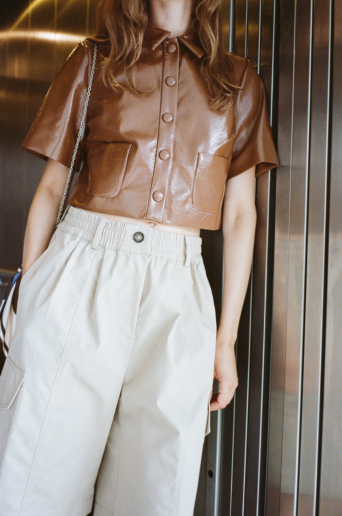 Close-up campaign styling of a glossy brown short-sleeved button-front jacket with large flap pockets and cream trousers.