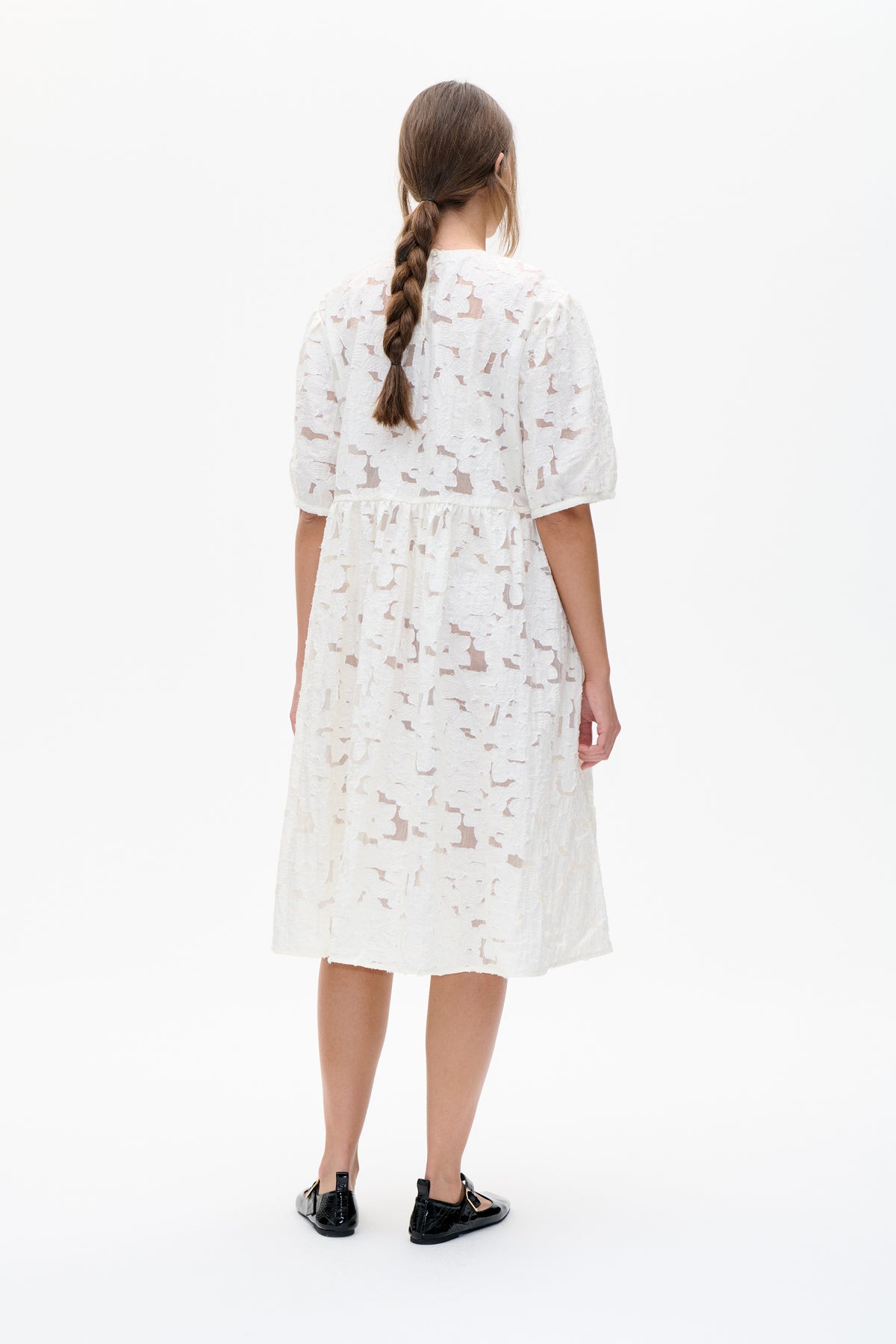 Back view of a white short-sleeved dress with sheer floral lace, a gathered waist and a tiered skirt.