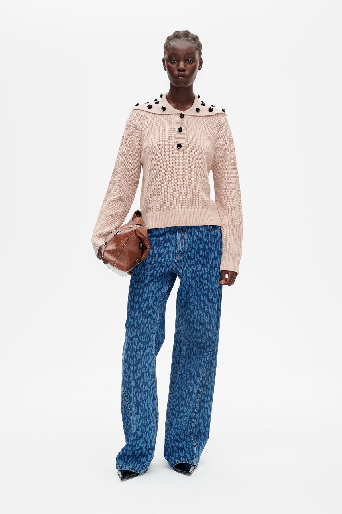 Model wearing a pale pink ribbed sweater with a wide collar and black floral embellishments, paired with blue patterned jeans.