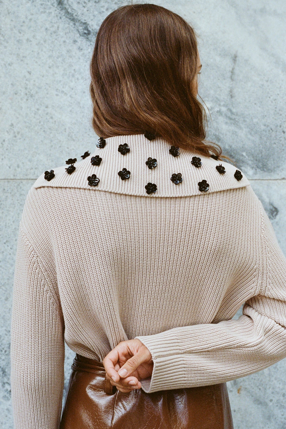Campaign close-up from behind of a cream textured knit sweater with black floral embellishments across the shoulders and a dark brown glossy garment.