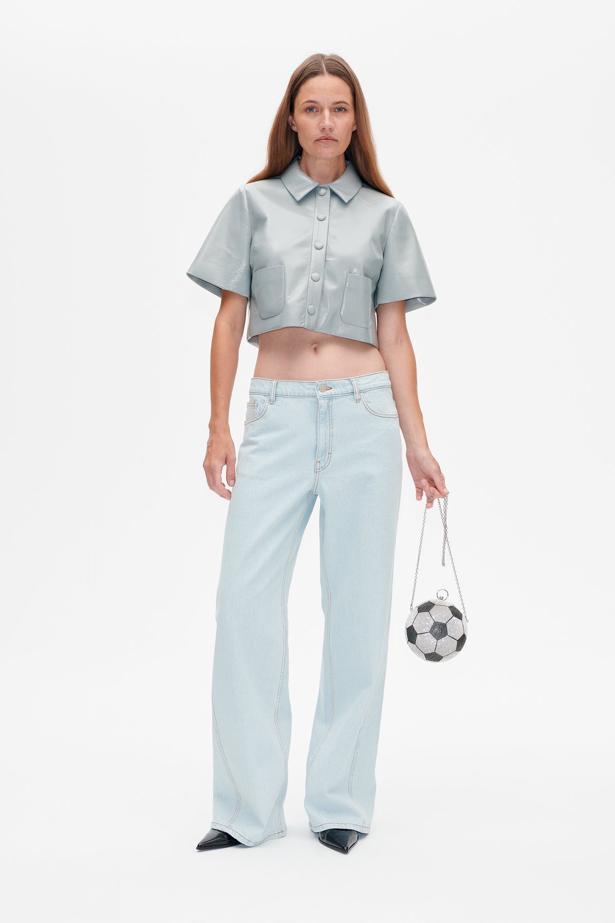 Ganzkörper-Studioaufnahme eines Models in einem hellblauen, glänzenden, kurz geschnittenen Hemd mit aufgesetzten Taschen, hellen, weit geschnittenen Jeans und einer fußballförmigen Tasche.