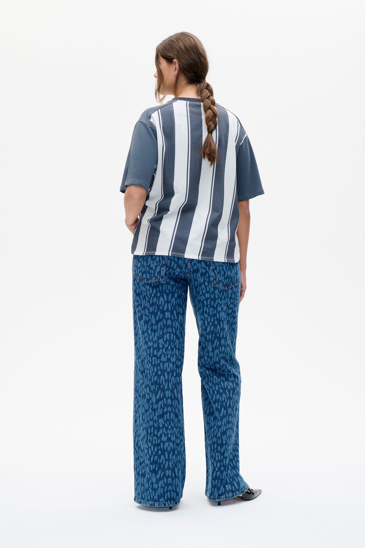 Rückansicht eines Models, das ein blau-weiß vertikal gestreiftes T‑Shirt mit kurzen Ärmeln und eine gemusterte, weit geschnittene blaue Jeans trägt.