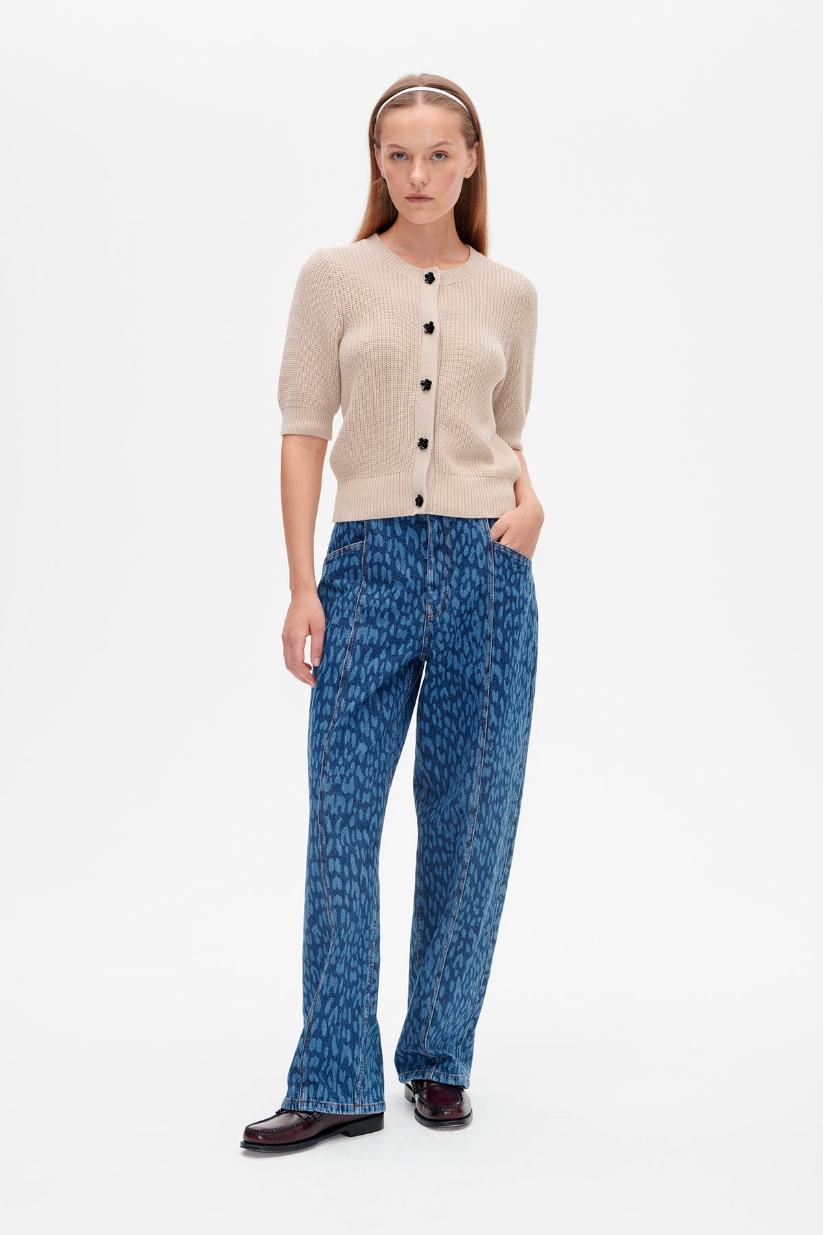 Model trägt einen cremefarbenen, kurzärmeligen Strickcardigan mit schwarzen, blumenförmigen Knöpfen und blau gemusterten Jeans, von vorne gezeigt.