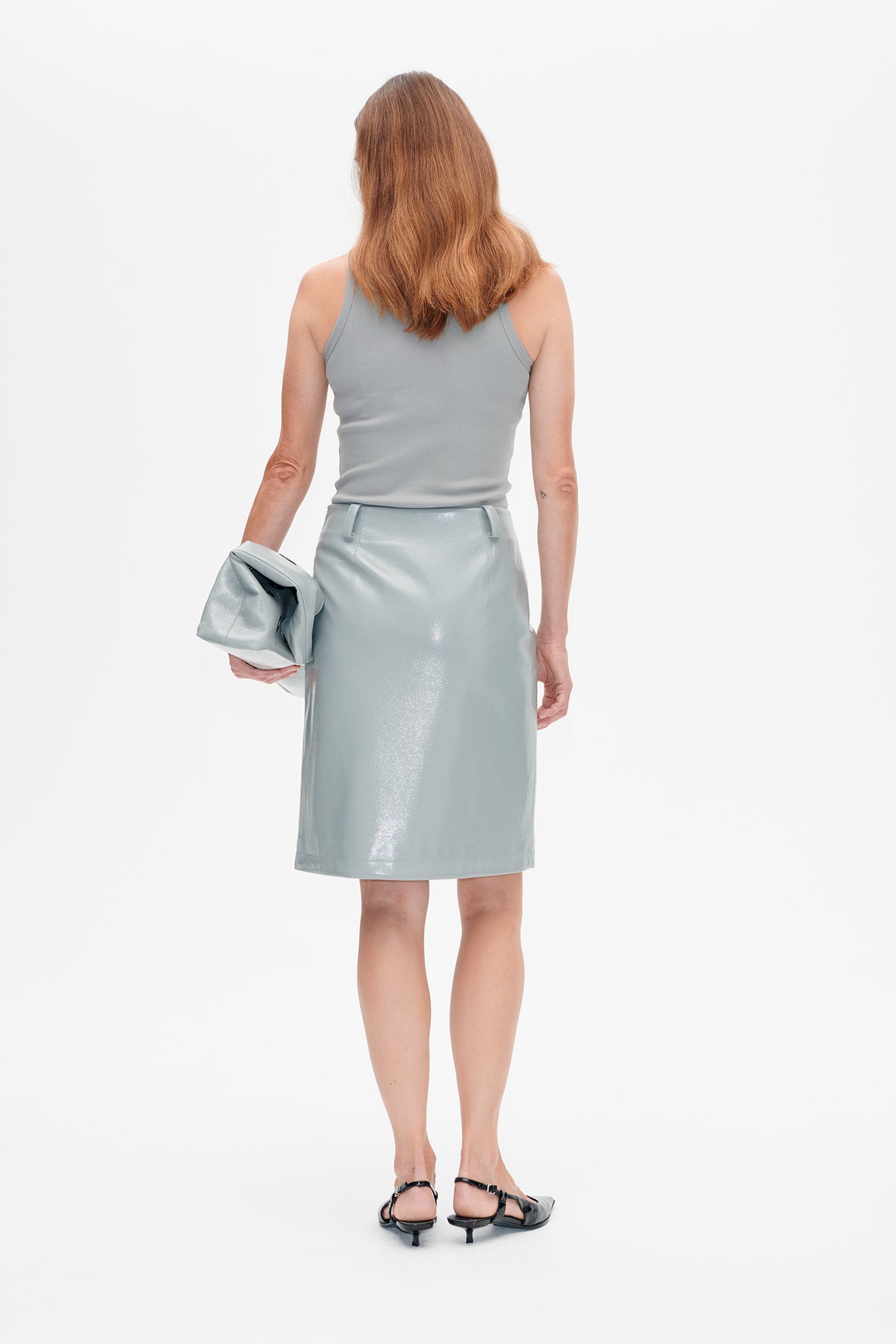 Back view of a model wearing a pale blue sleeveless top and glossy matching knee-length skirt, holding a clutch.