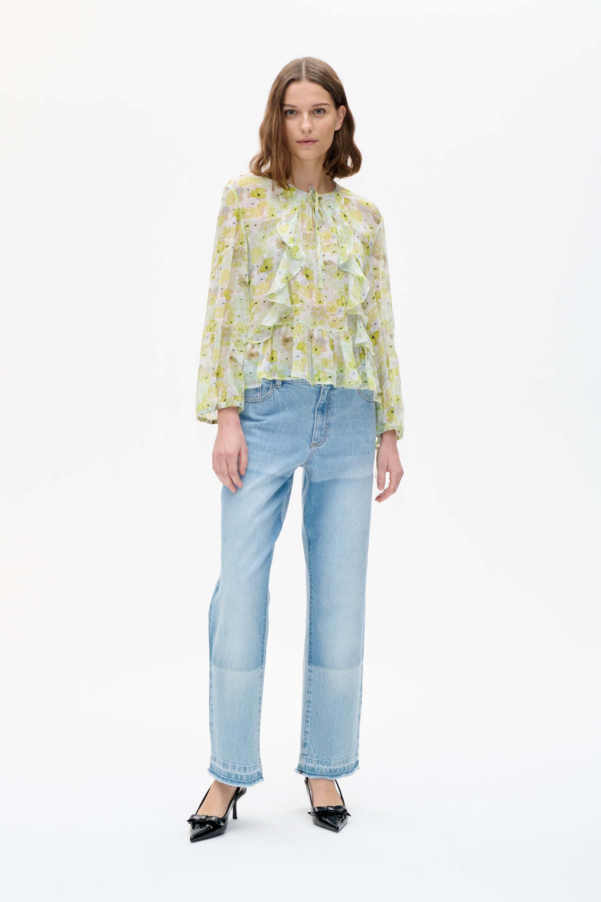 Vooraanzicht van een bloemige blouse in lichtgeel en groen met lange mouwen, een strik bij de hals en vallende ruches aan de voorkant, gedragen met lichtgewassen jeans.