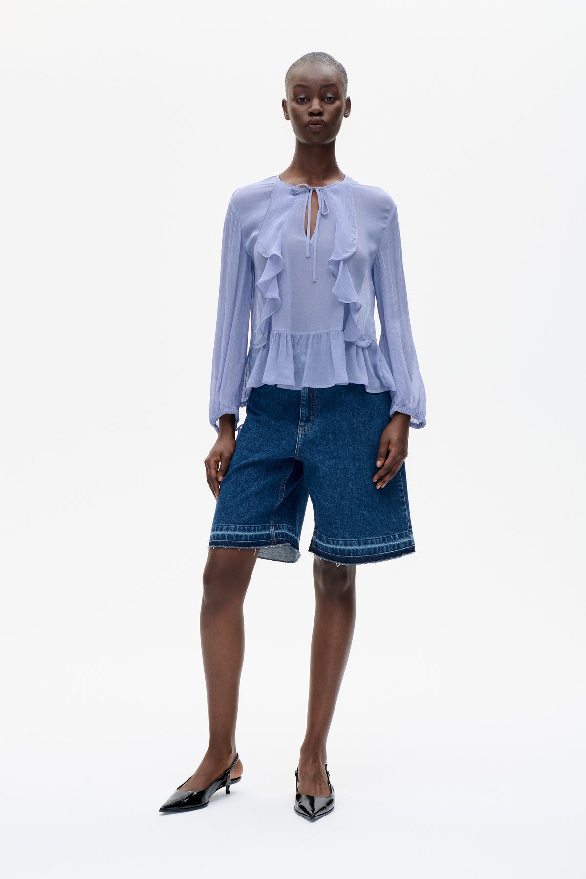 Model draagt een licht lavendelkleurige blouse met lange mouwen, een striksluiting bij de hals en vallende ruches, gecombineerd met donkere denimshorts.