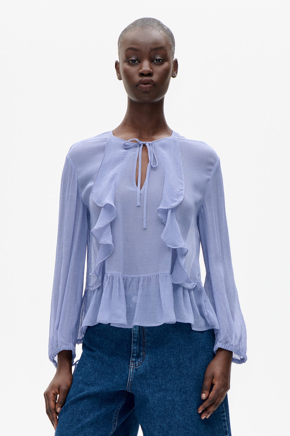 Front view of a pale blue long-sleeved blouse with a tie keyhole neckline, cascading ruffles and elastic cuffs, paired with dark jeans.