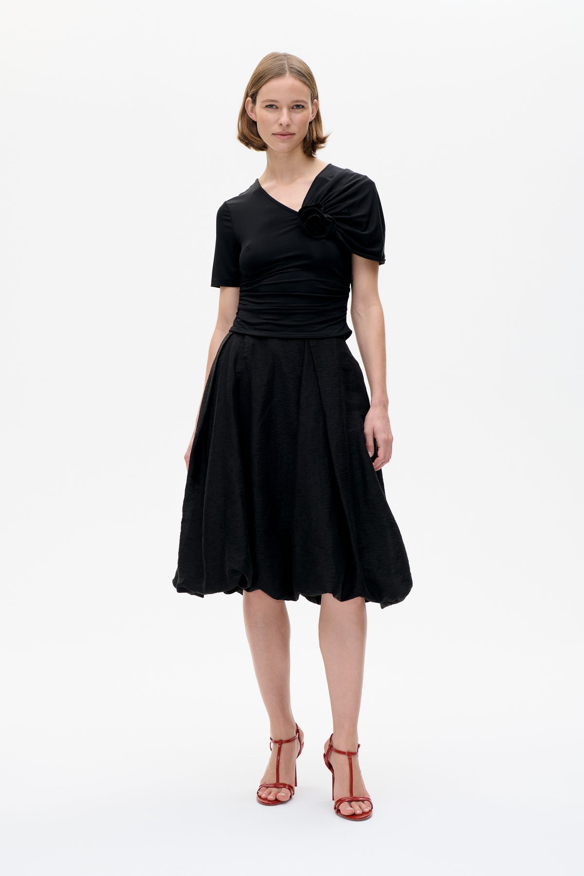 Front view of a model wearing a black asymmetrical top with a sculptural shoulder rosette and ruched waist, paired with a voluminous black skirt.