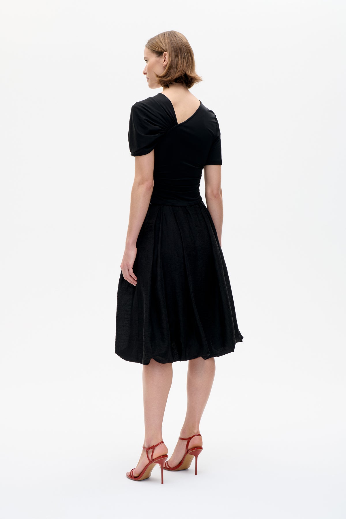 Model viewed from behind wearing a black short-sleeved top with an asymmetrical open back and a full, textured black skirt.